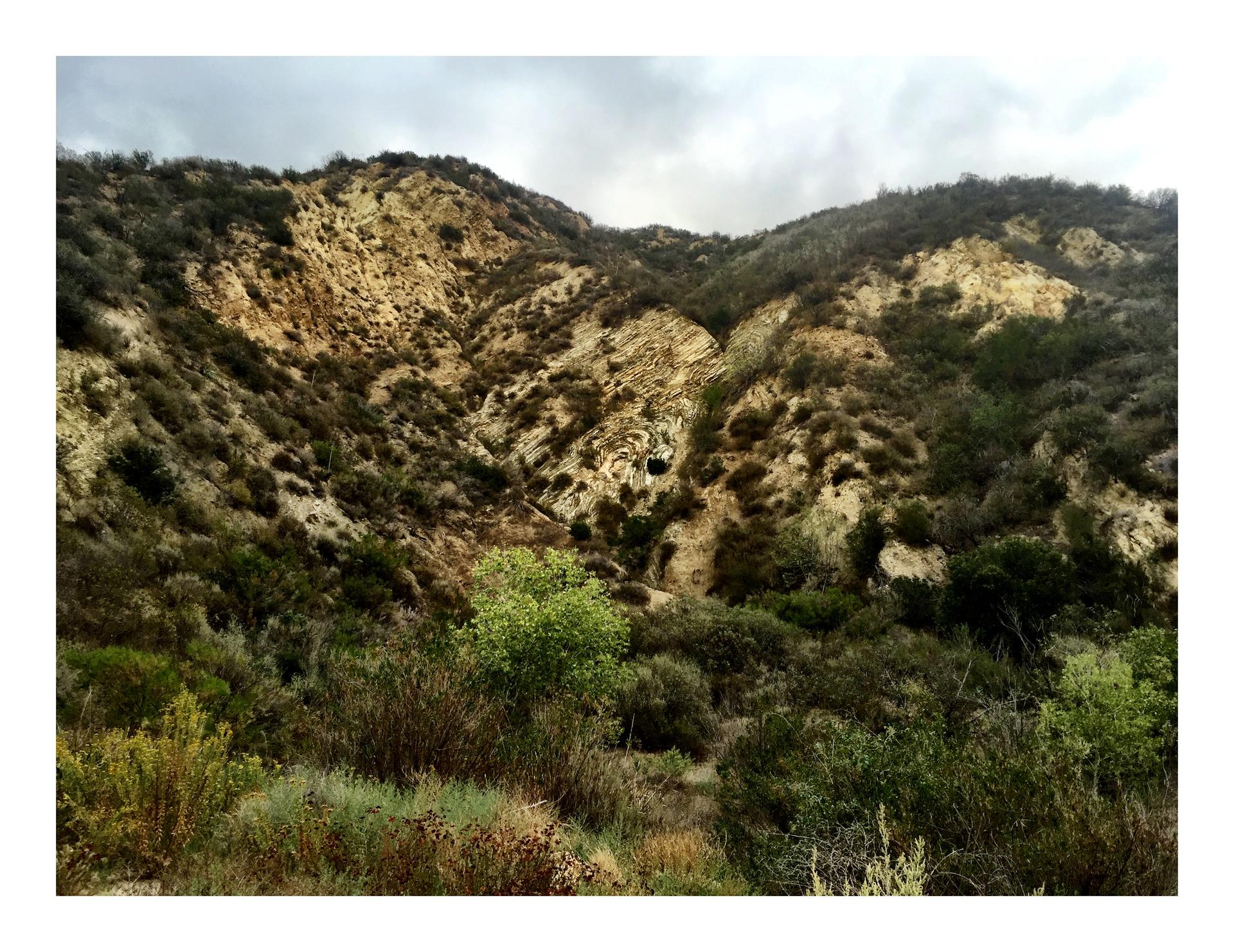 Steep, eroded hillside with green brush and exposed rock under an overcast sky.