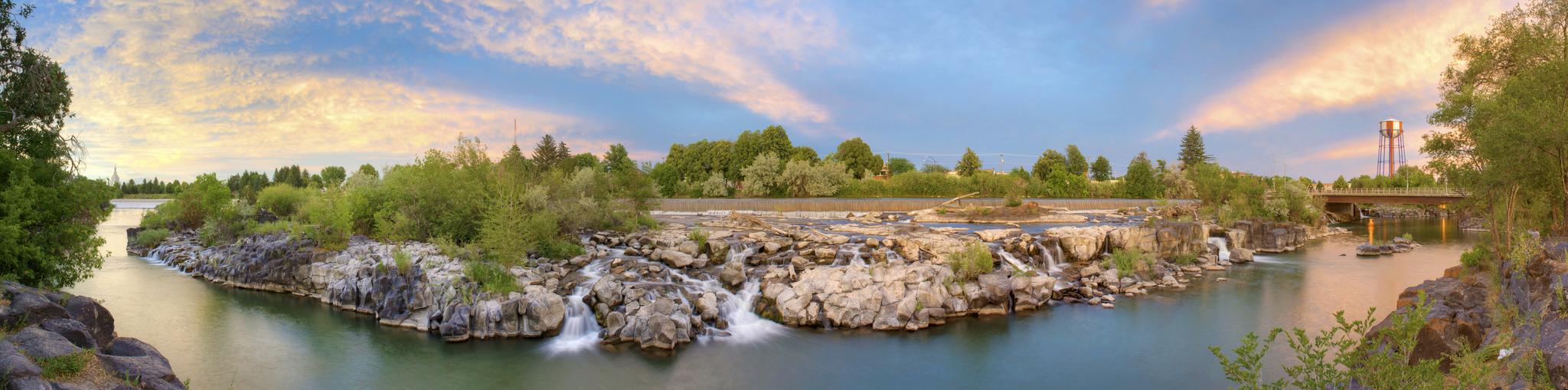 Panoramic river view with waterfalls, green trees, and a vibrant sunset sky.