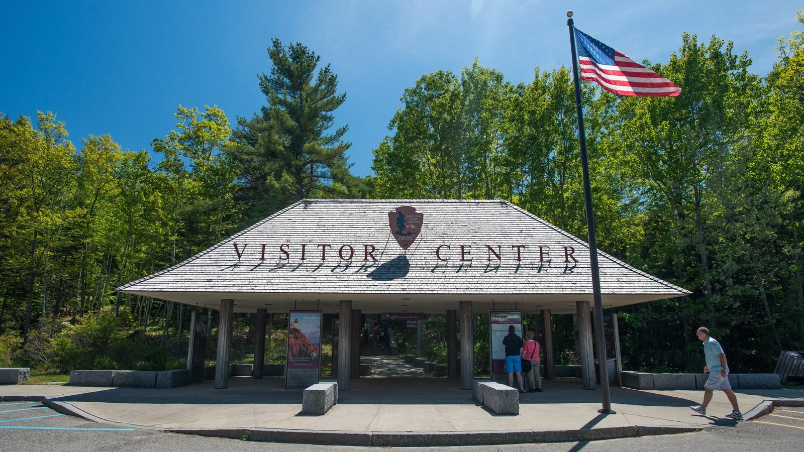 People walk near a visitor center building with a flag on a sunny day.
