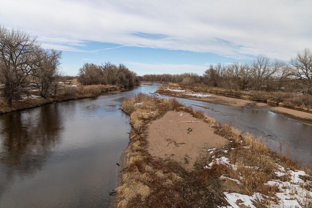 A wide river flows around a sandy island with bare trees and snow.