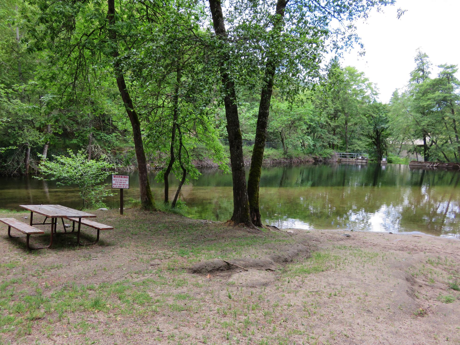 A picnic table on a sandy bank next to a calm river and trees.