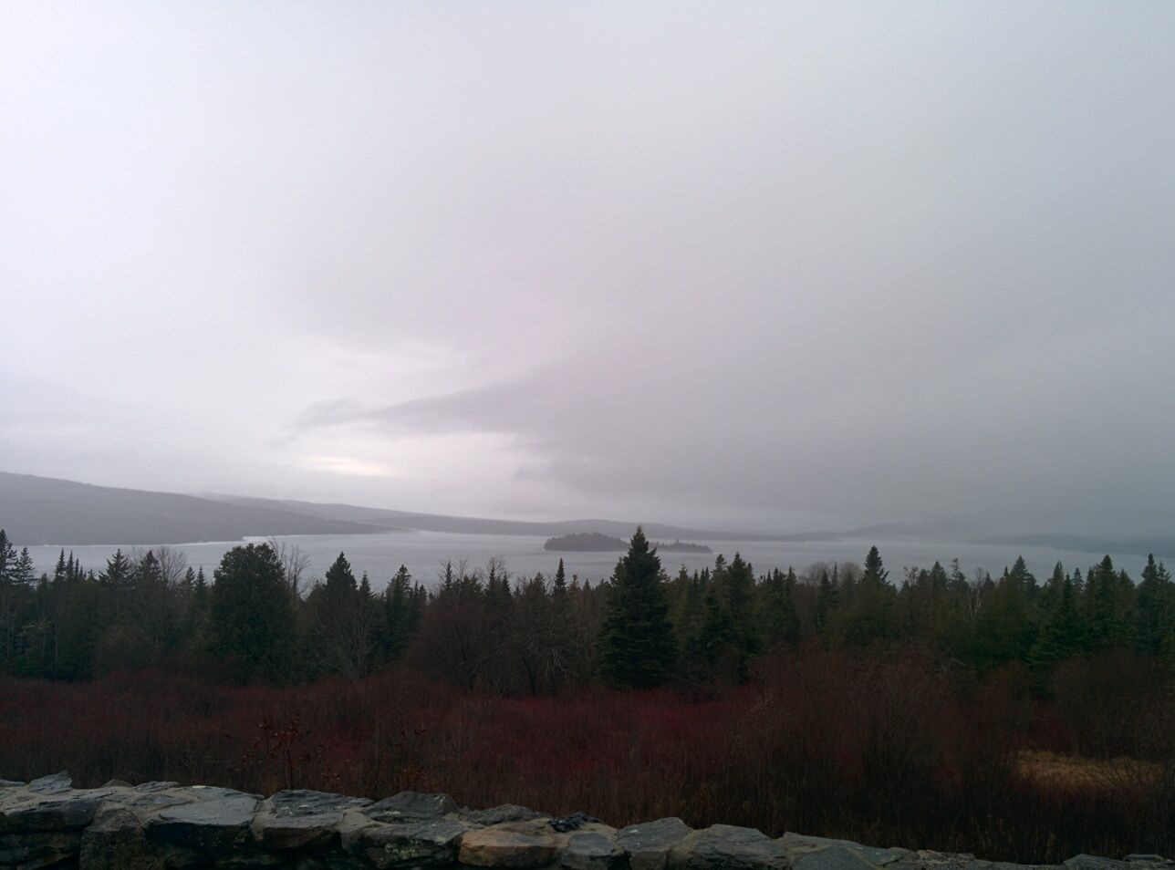 Overcast landscape with a large lake, dense forest, and distant misty mountains. - Rangeley Lake loop