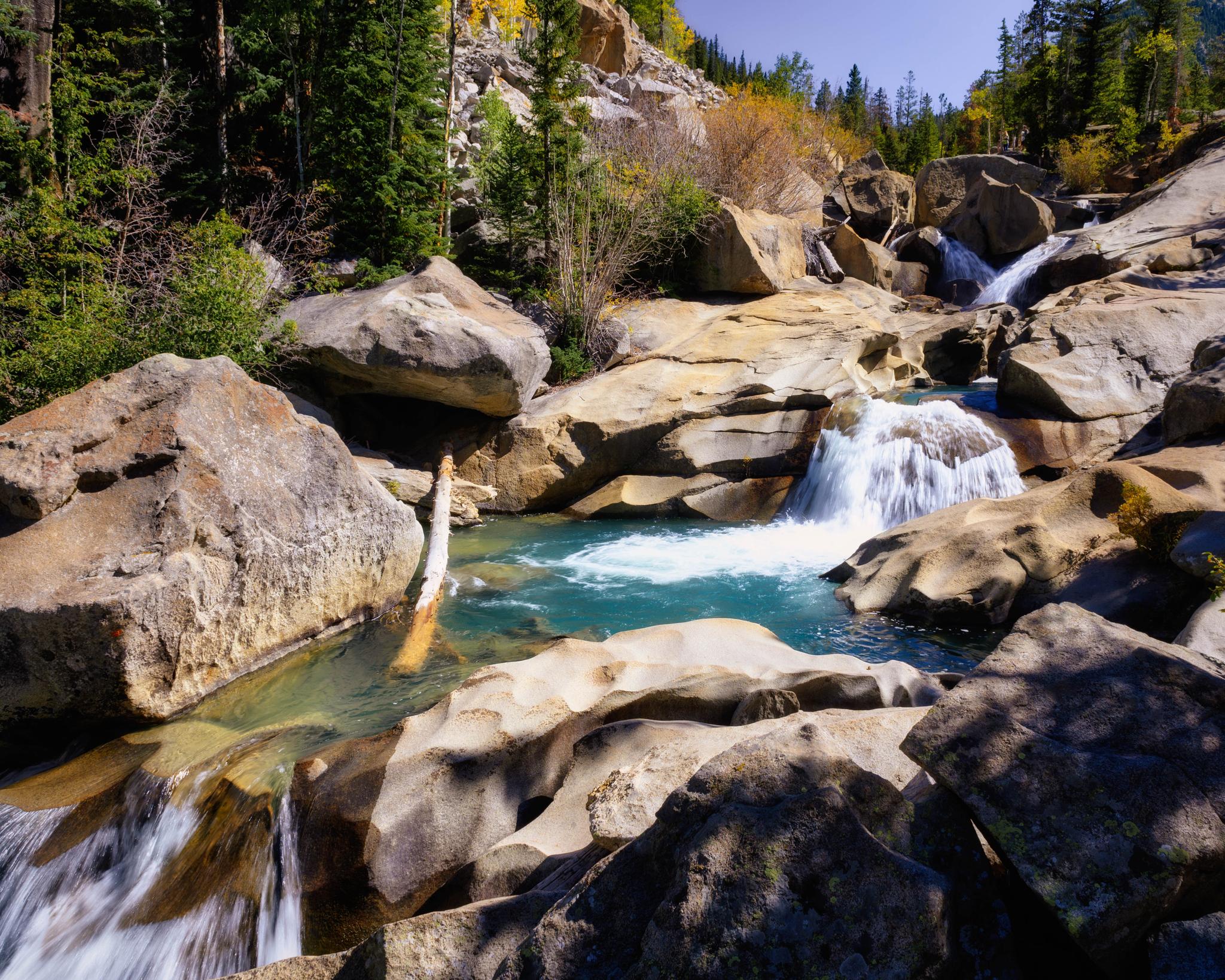 Clear blue water cascades over large boulders in a sunny, rocky river.