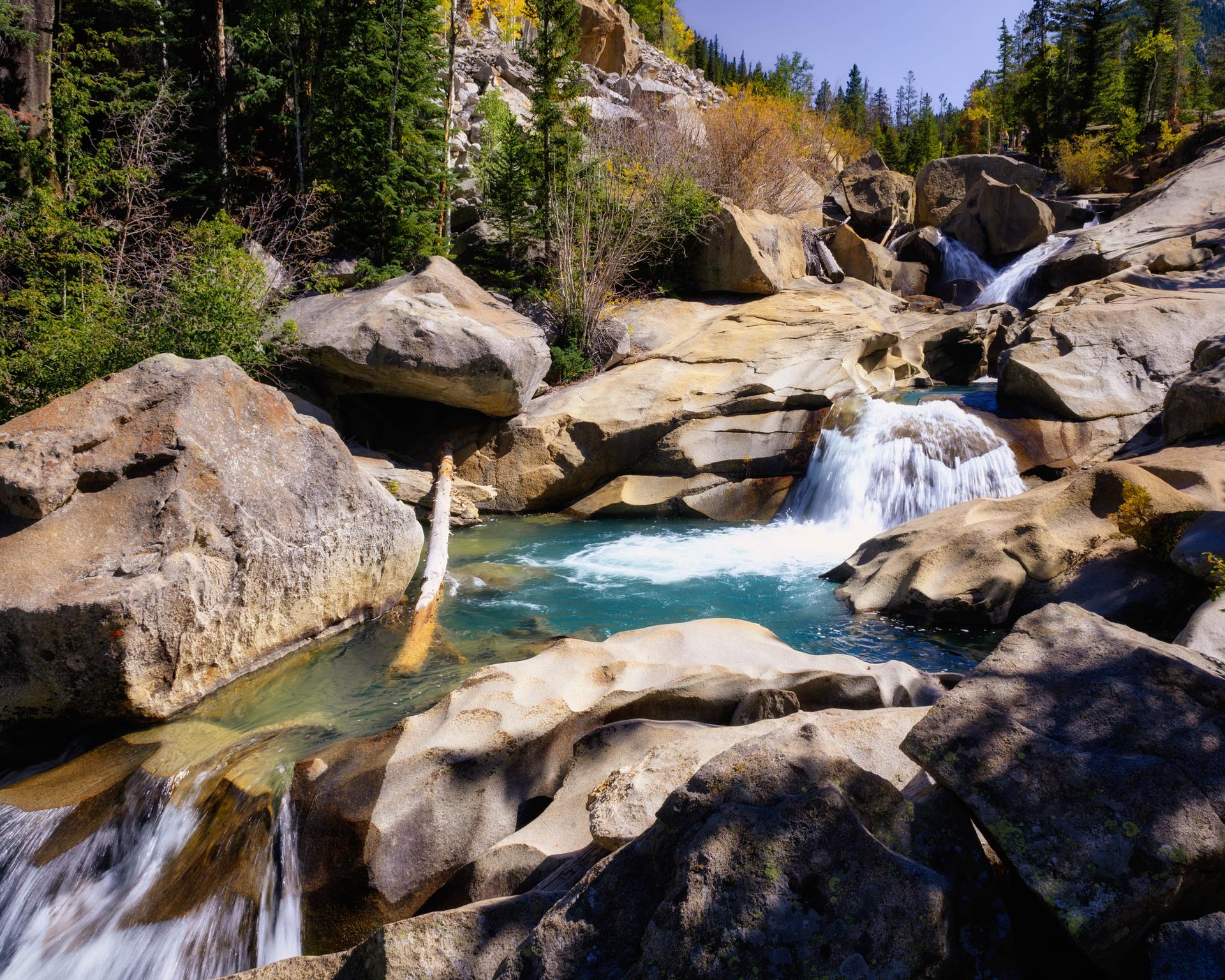 Clear blue water cascades over large boulders in a sunny, rocky river.