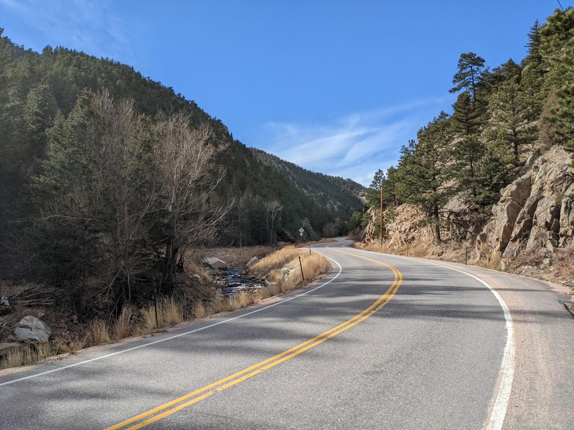 A winding paved road through a forested canyon with a stream.