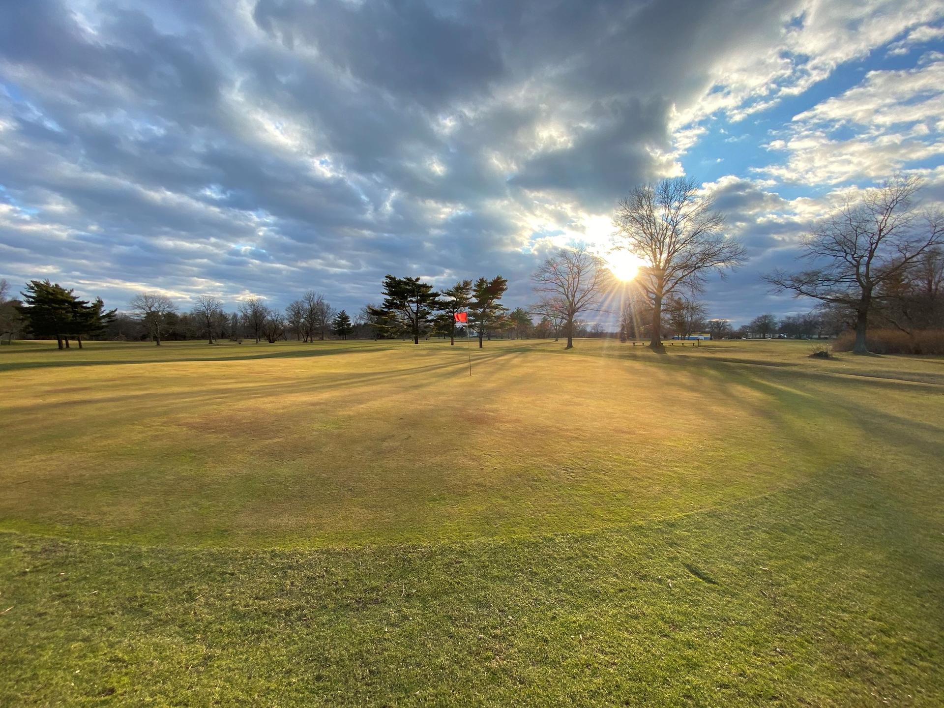 Golden hour sunburst over a green golf course with scattered trees and cloudy sky.