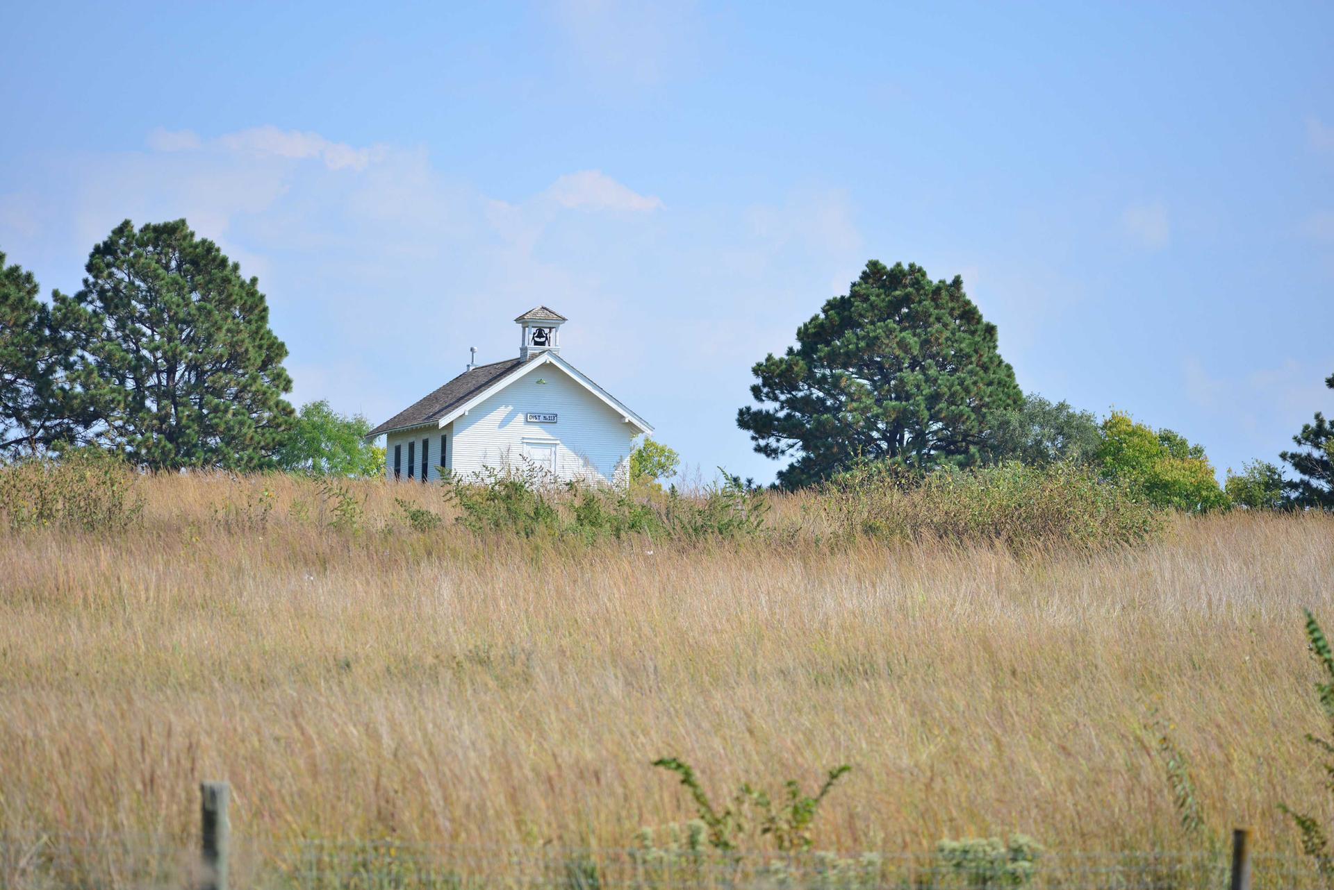 White building with bell tower on a grassy hill under a bright sky.