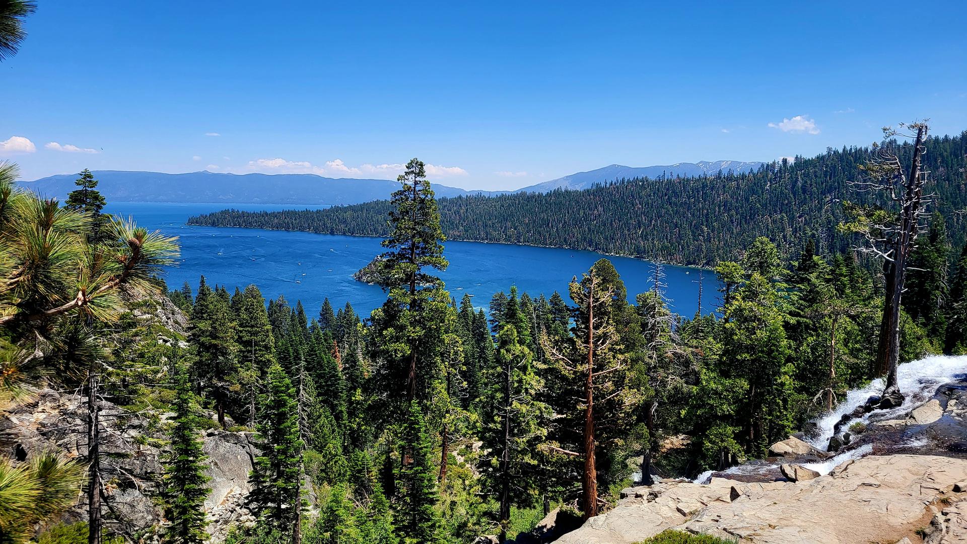 Wide blue lake surrounded by dense pine forests and mountains, small waterfall.