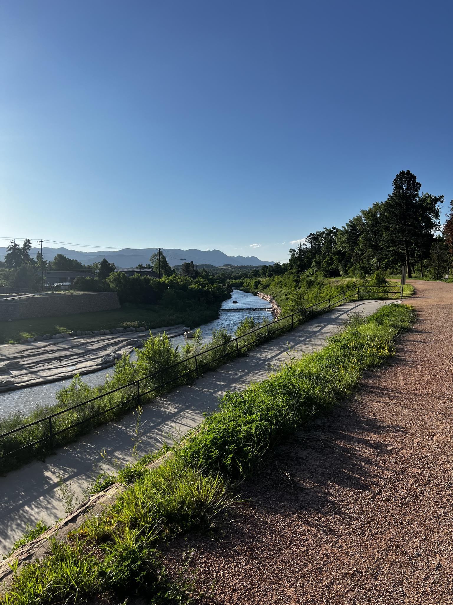 Sunny view of a red path, river with concrete banks, and distant mountains.
