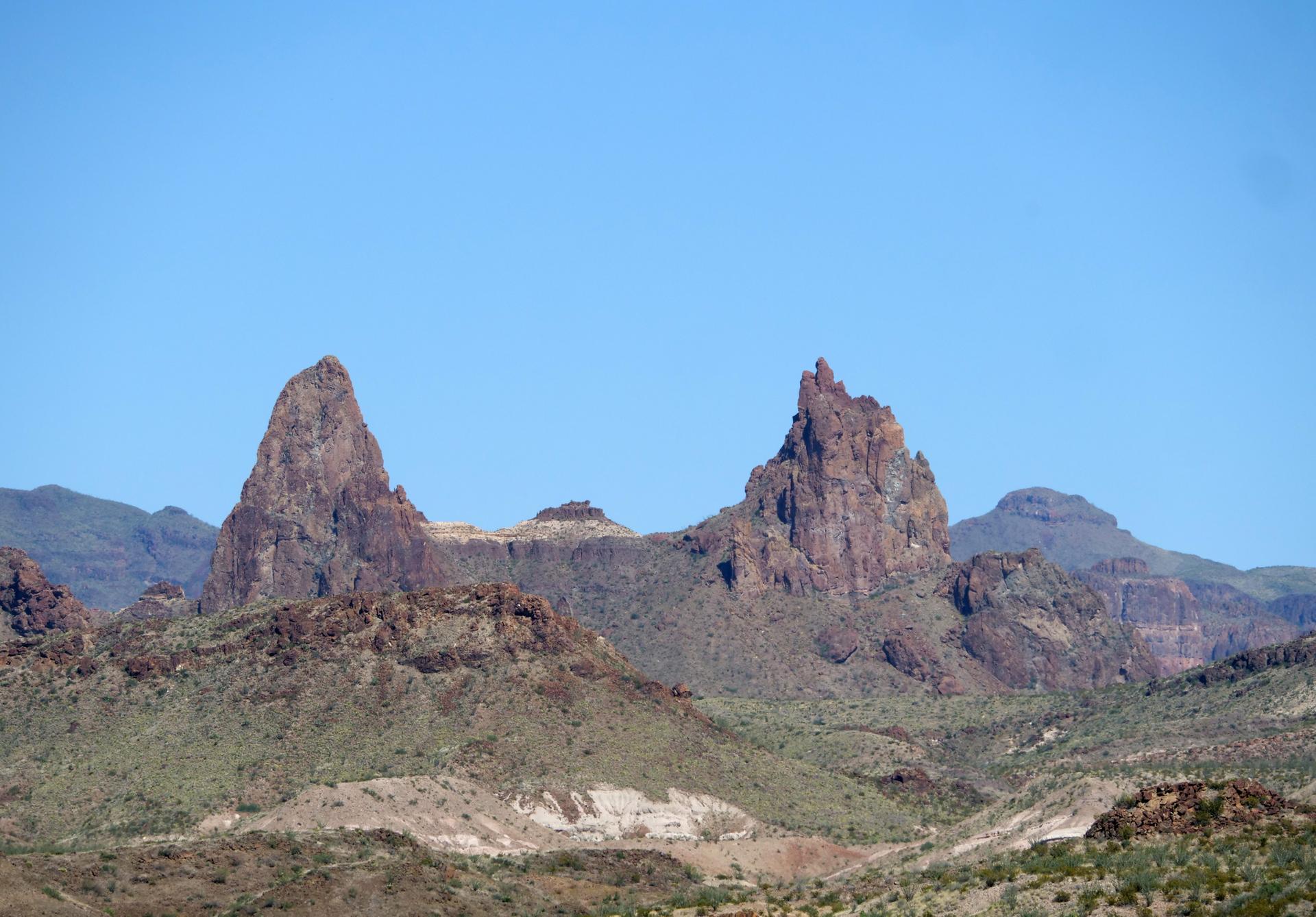Two prominent rocky peaks rise from a desert mountain range, clear sky.
