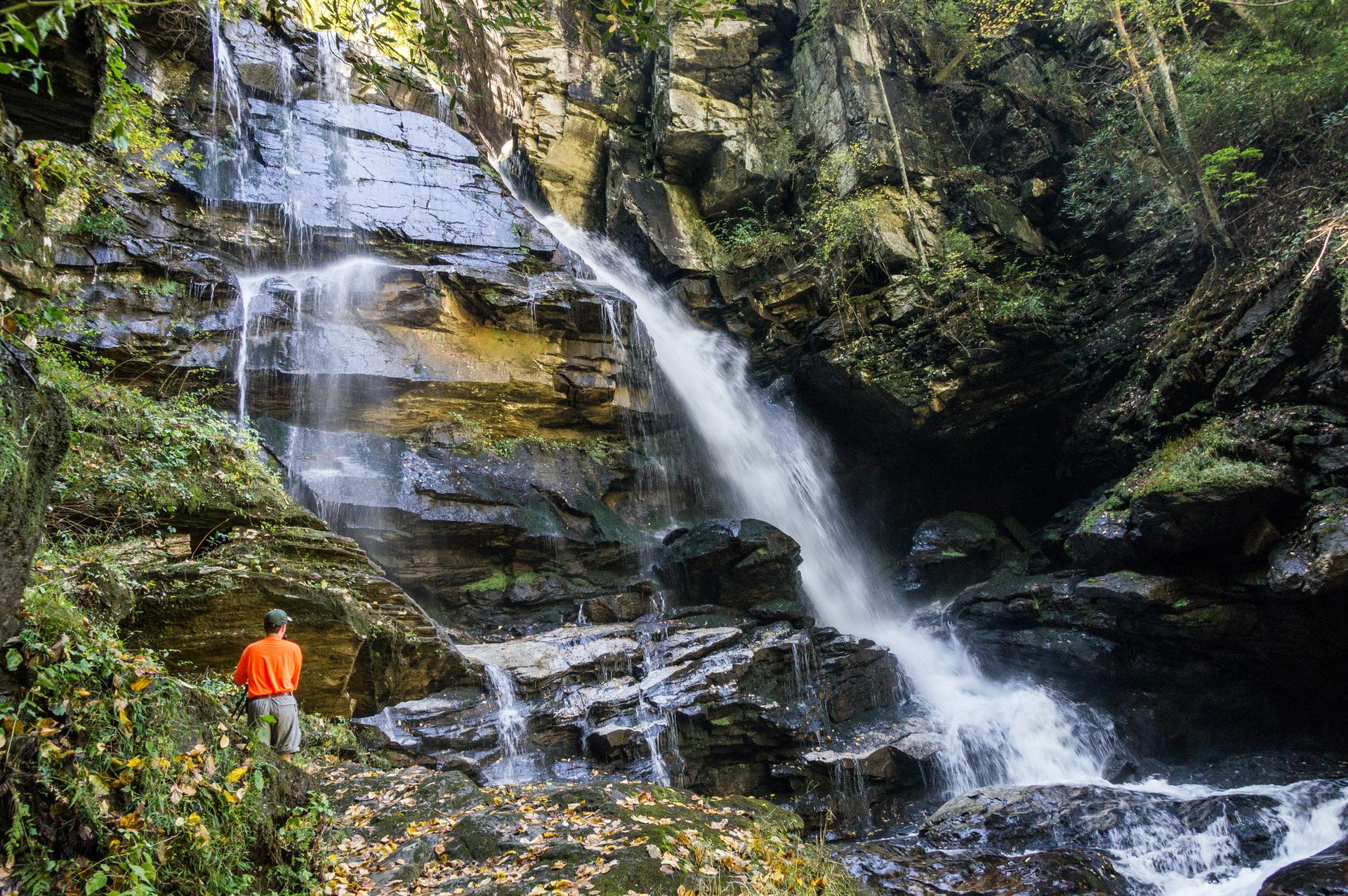 A person in an orange shirt stands admiring a multi-tiered waterfall in a lush forest.