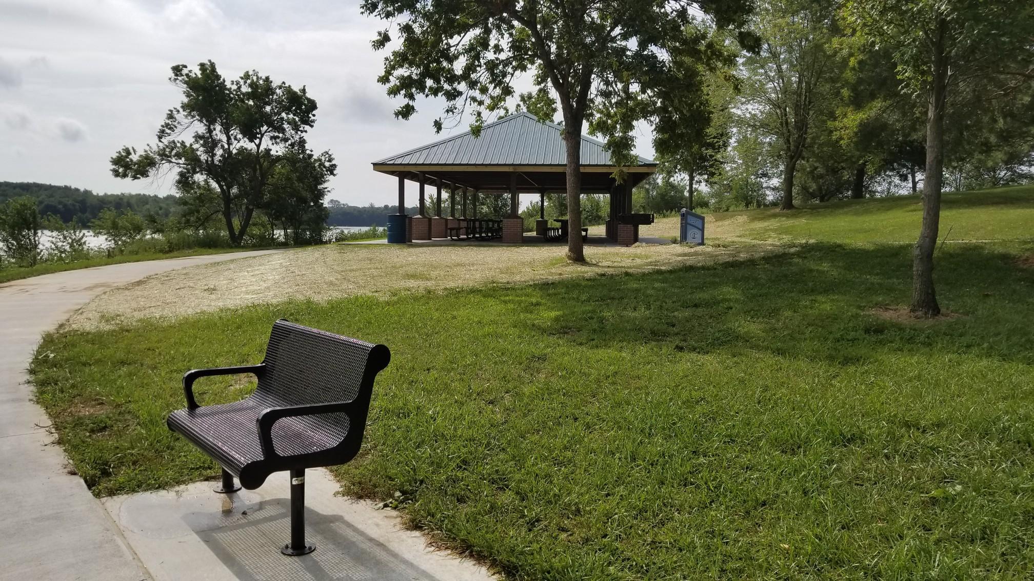 Park bench by a paved path, with a picnic shelter and lake.