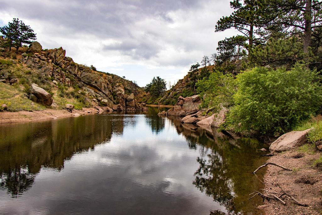 Calm lake reflecting cloudy sky, surrounded by rocky banks and pine trees.