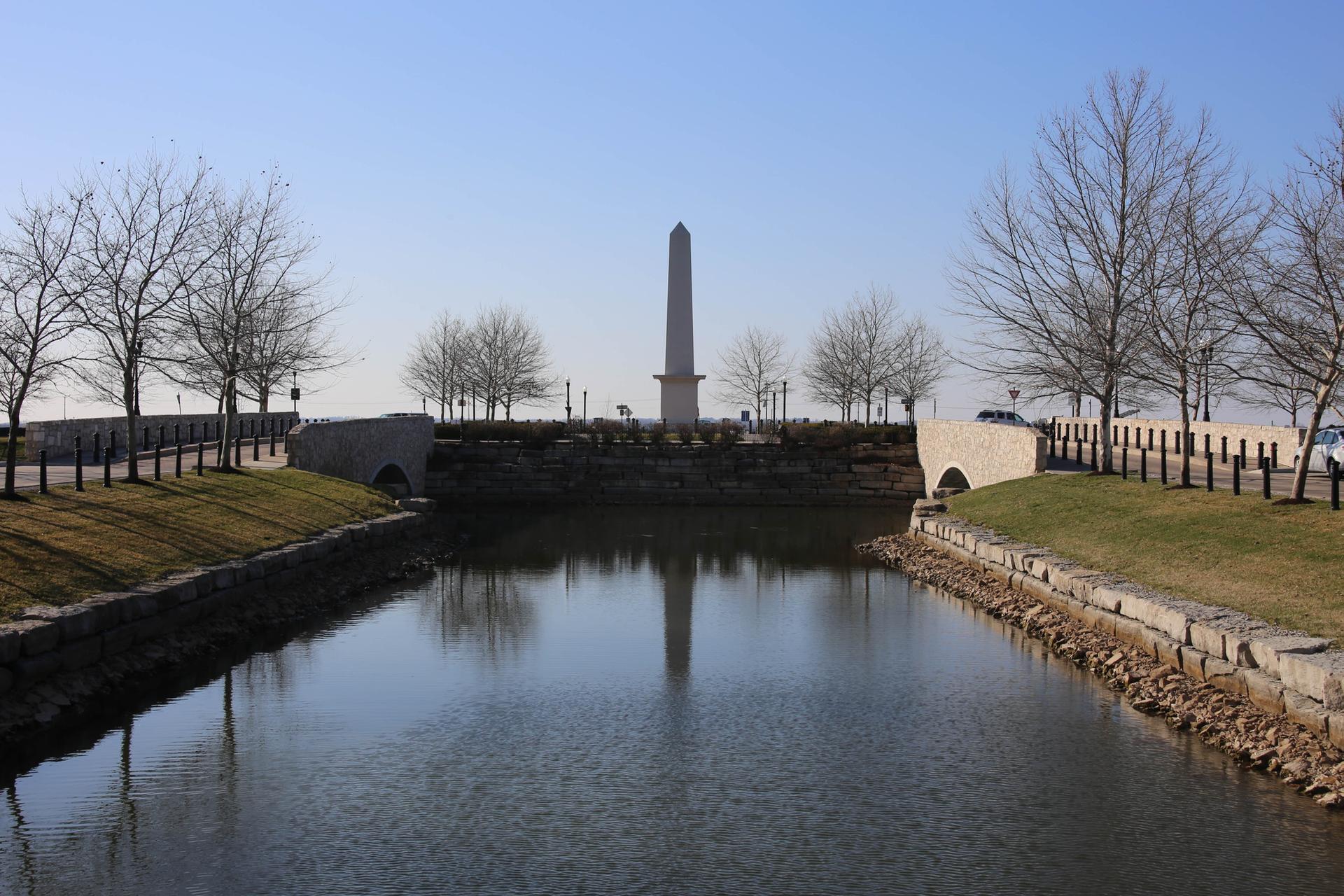 A canal with an obelisk monument, bare trees, and stone bridges under clear skies.