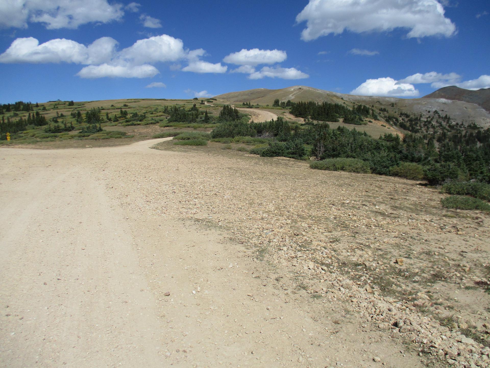 A dirt road forks through sunny, rocky, mountainous terrain under a blue sky.