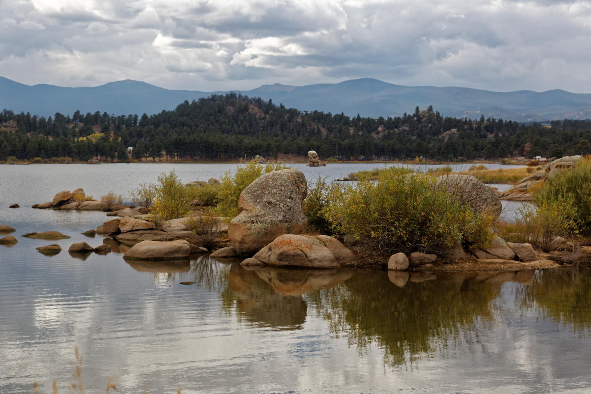 Rocky lake shore with reflections, forested hills, and distant mountains under cloudy sky.