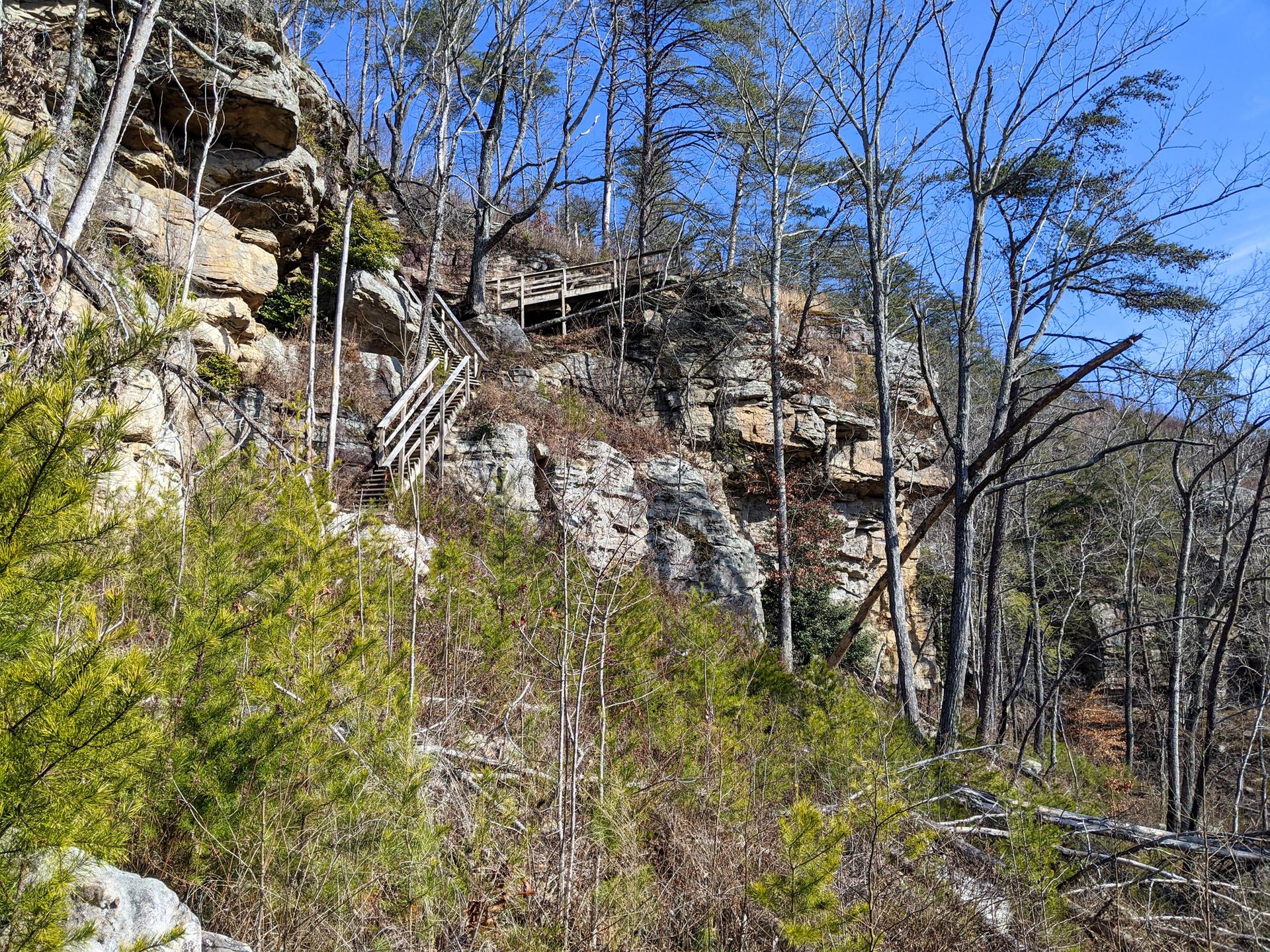 Wooden stairs and boardwalk ascend a rocky cliffside under a clear sky.