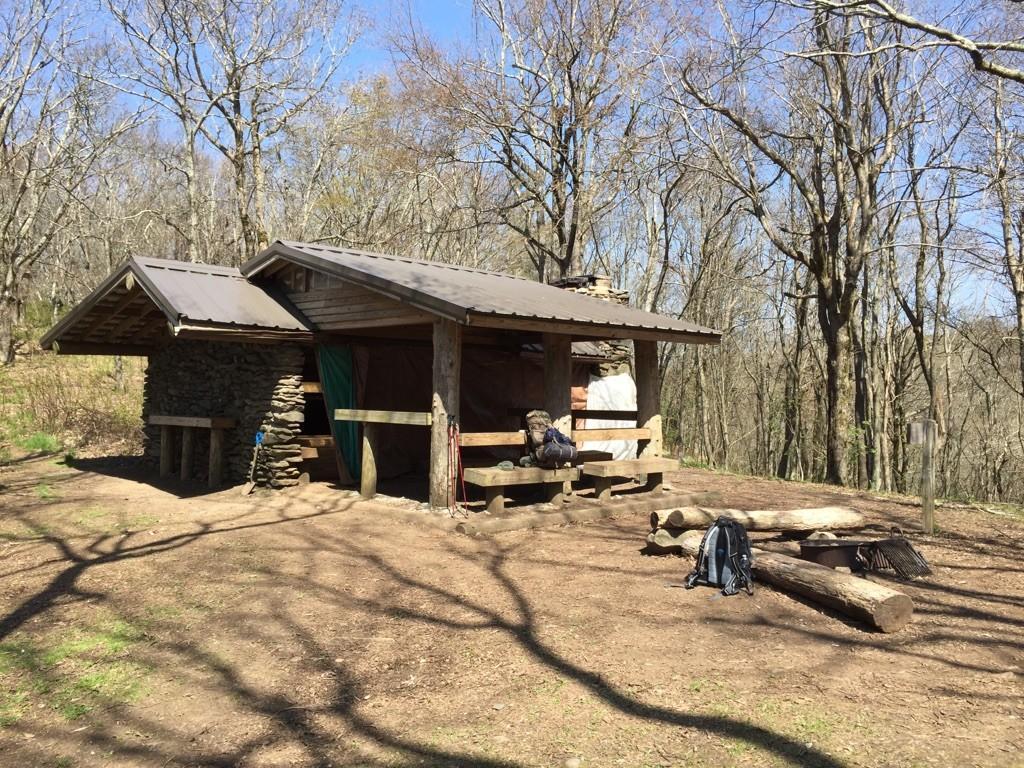Rustic wooden shelter with backpack in sunny, bare forest.