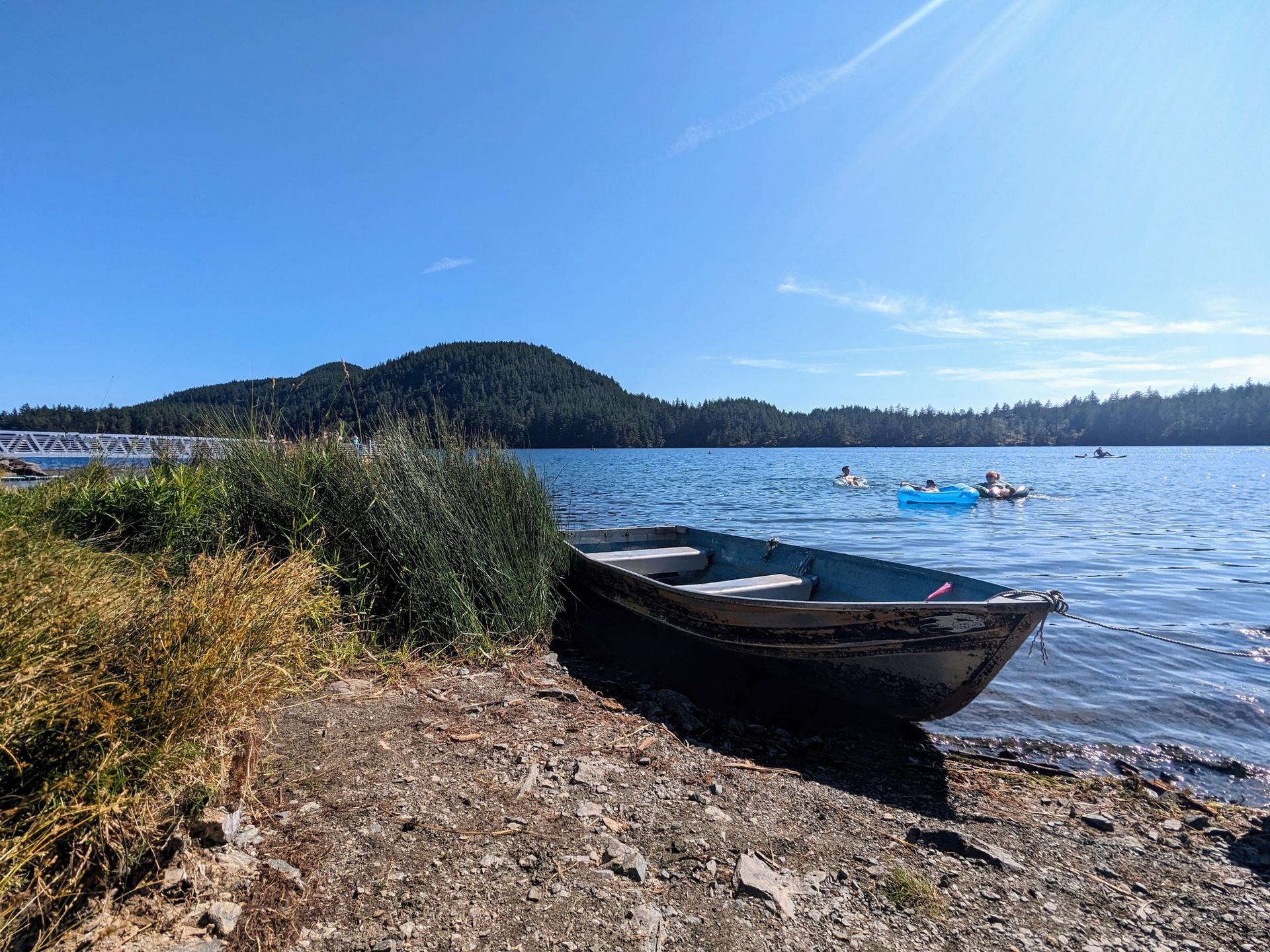 Small boat on a lake shore with forested hills and swimmers in bright sunlight.