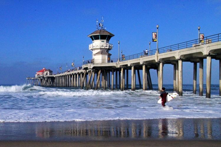 Surfer walking into ocean waves near a large pier with a white tower. Bright day.