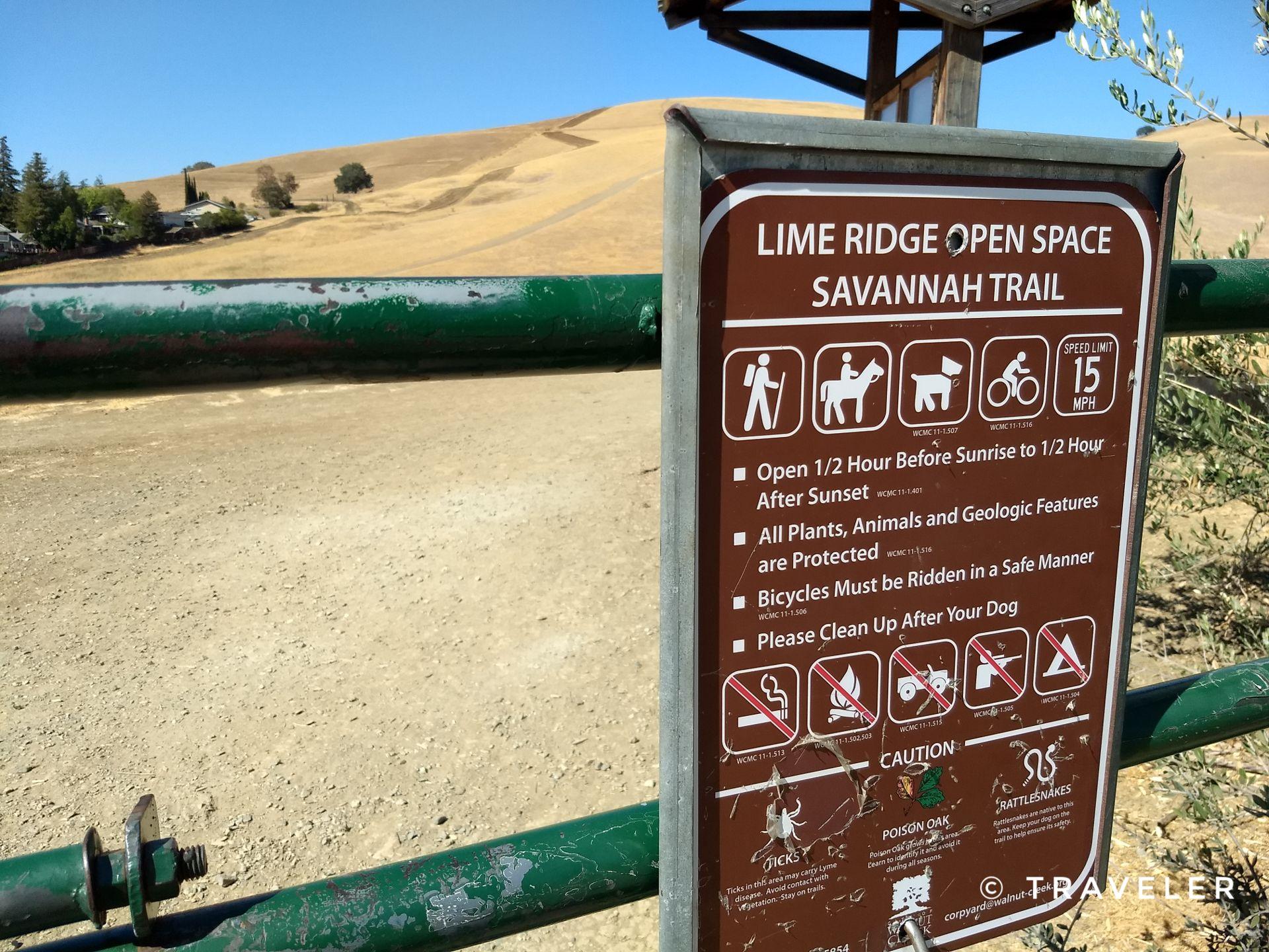 A trail sign on a green gate with dry golden hills under a bright blue sky.