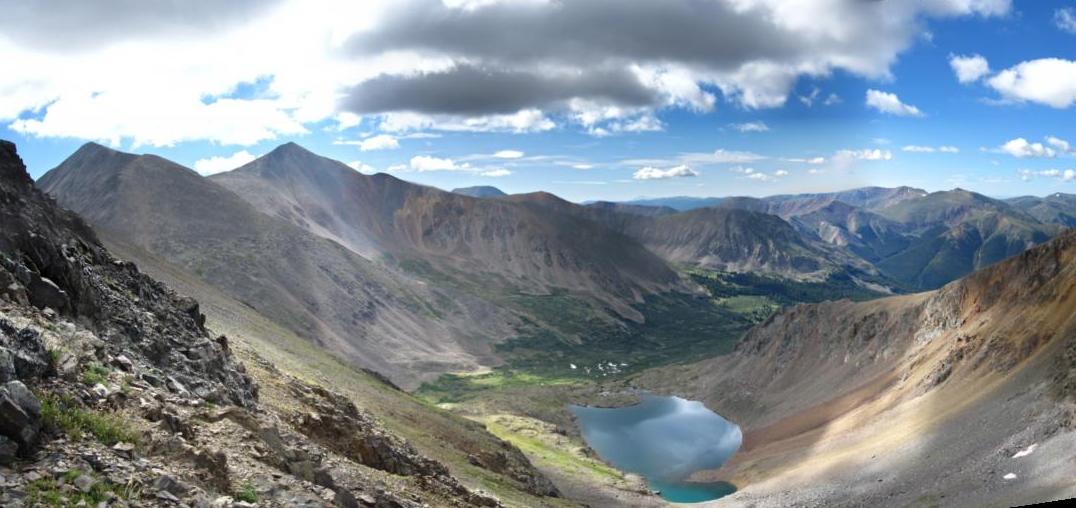 Panoramic view of rugged mountains, a valley, and a blue lake under a cloudy sky.