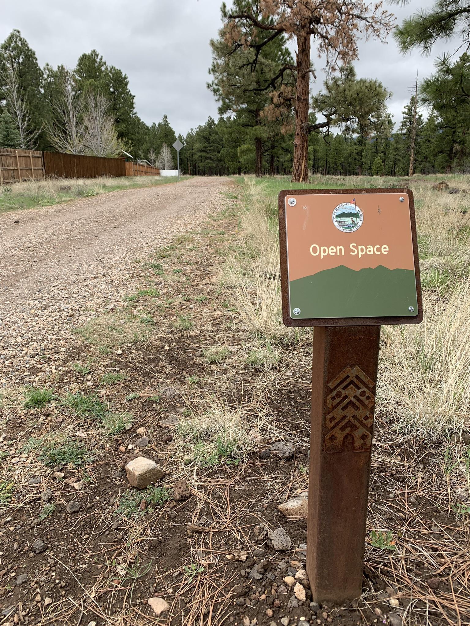 Overcast day, "Open Space" sign on a dirt path, surrounded by pine trees.