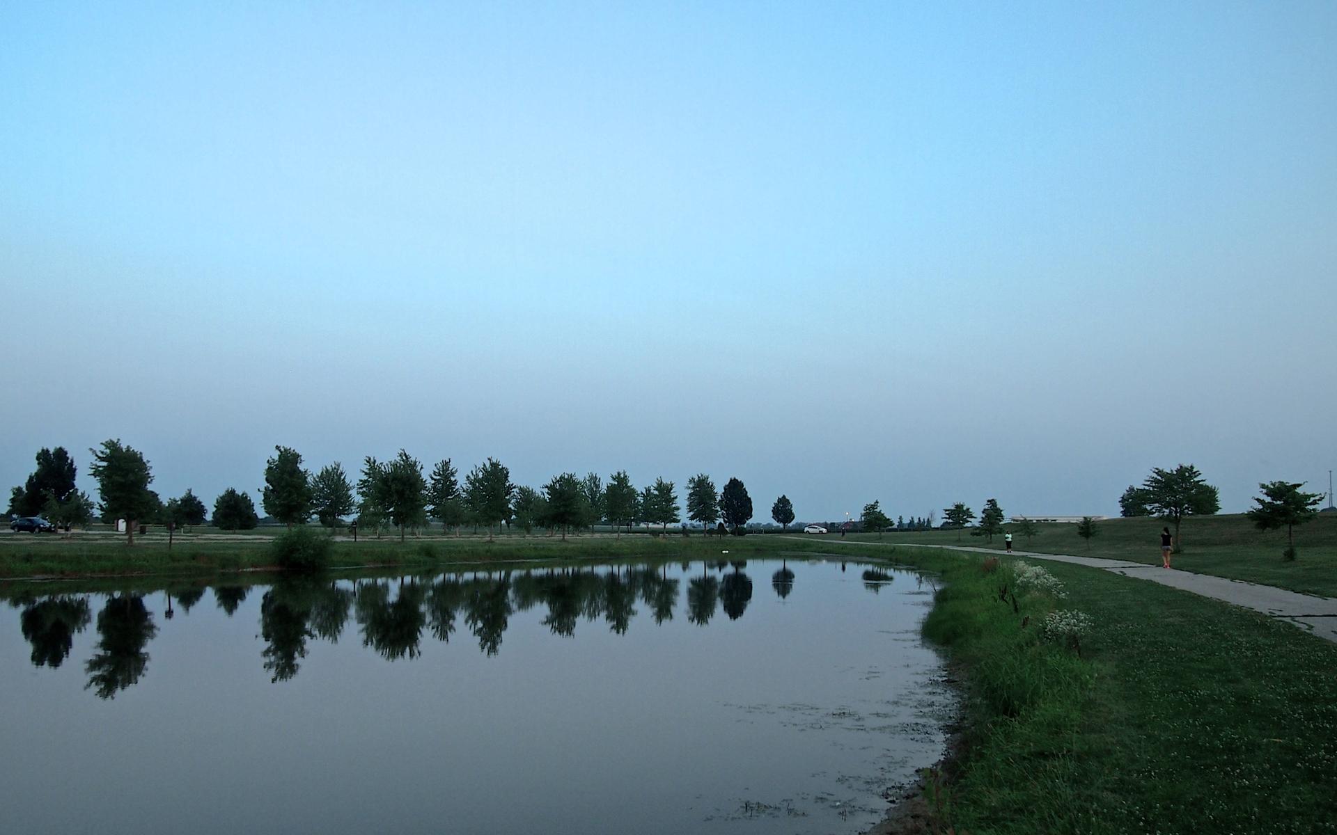 A calm pond reflects trees and a soft blue sky at dusk, with a path.