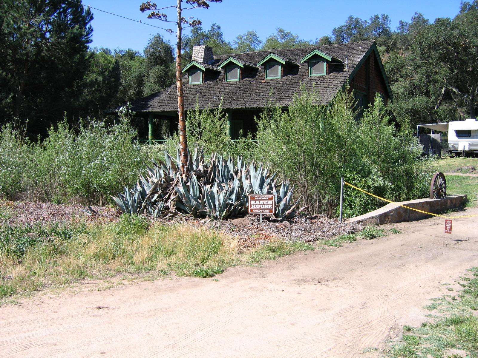 A rustic house behind agave plants and a dirt road on a sunny day.