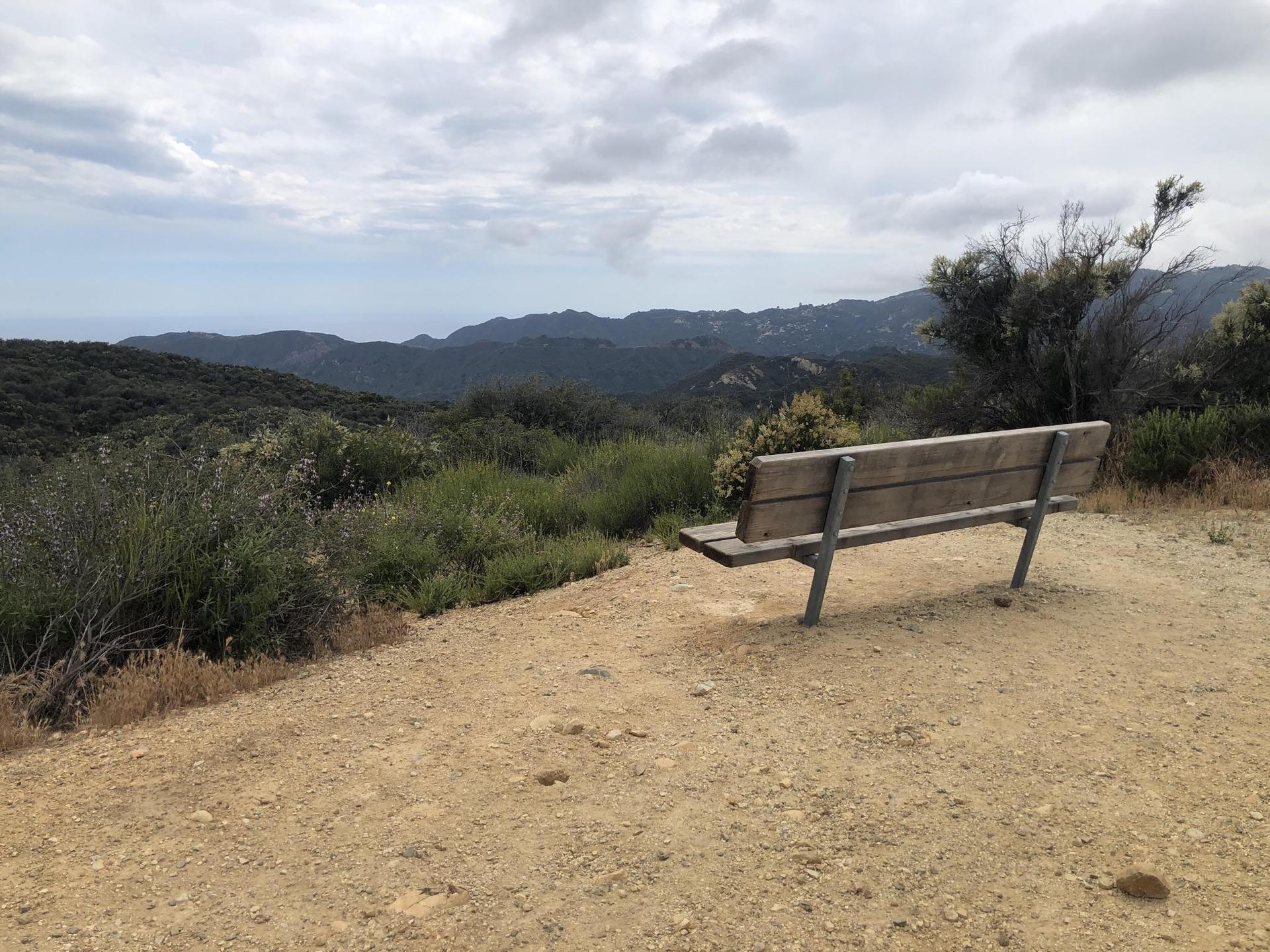 Empty wooden bench on a dirt clearing overlooking cloudy mountains and green hills.
