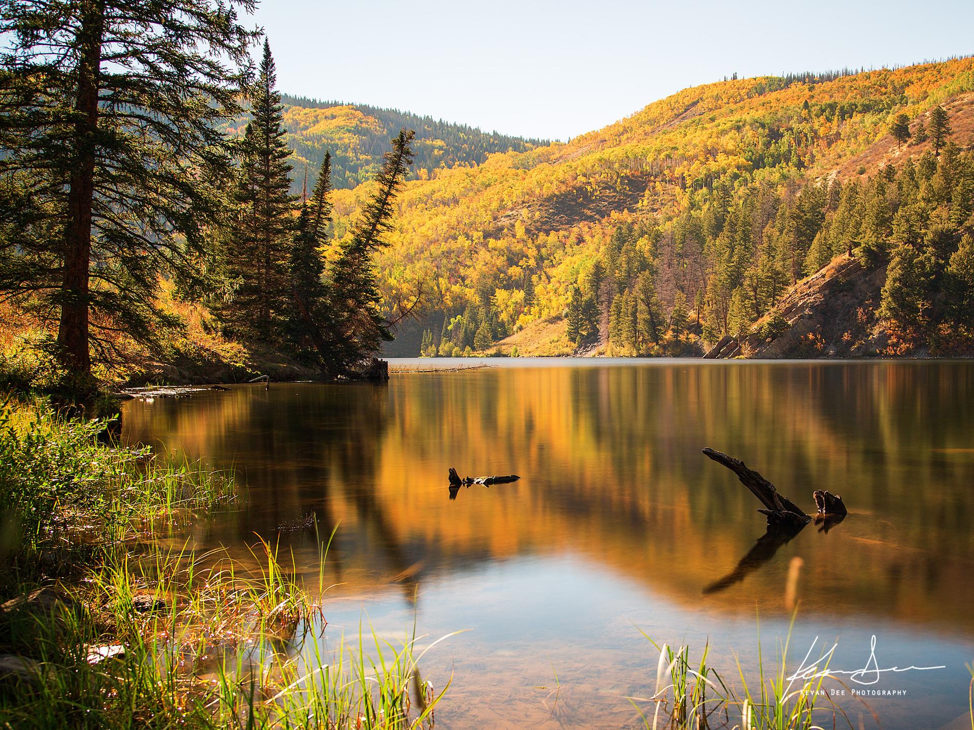 Calm lake reflecting vibrant autumn trees under bright sunlight.