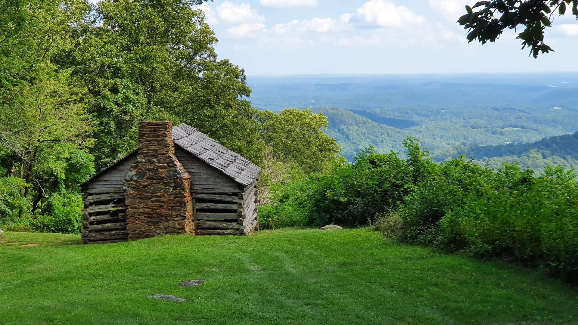 A rustic log cabin with a stone chimney overlooks hazy green mountains.