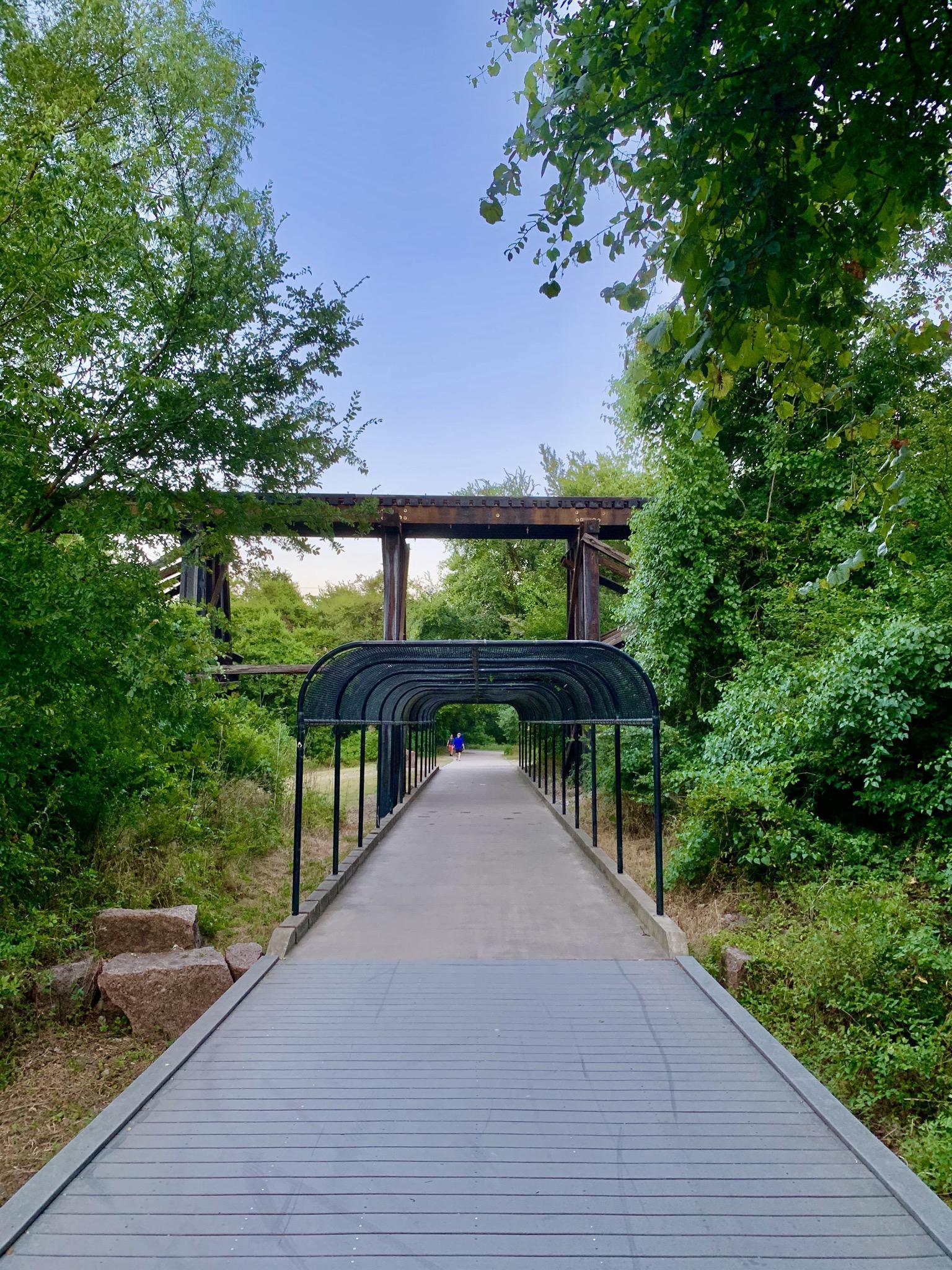 Covered walkway leading to a wooden bridge surrounded by green trees.