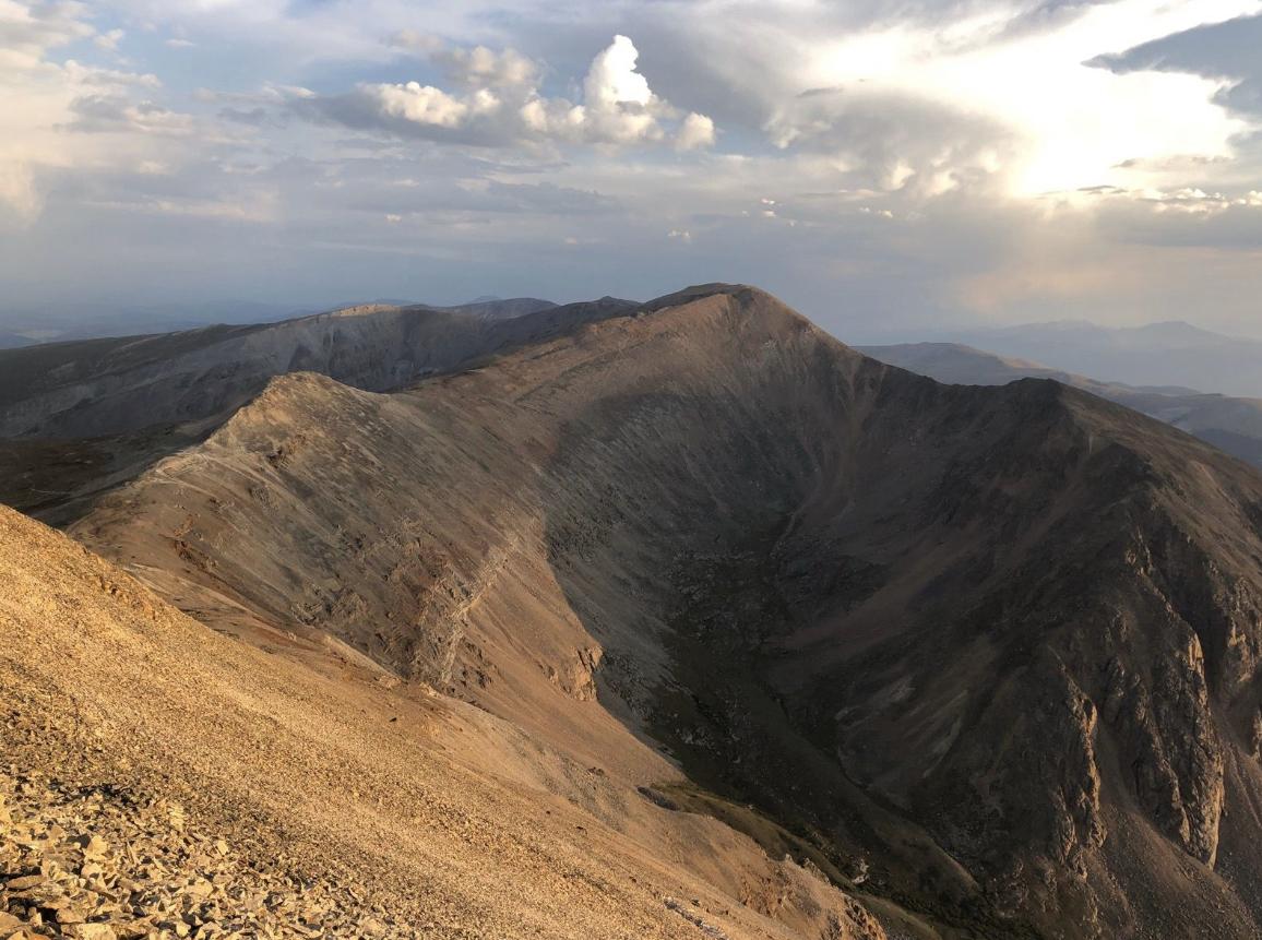 A dramatic, rugged mountain range with deep valleys under a cloudy, golden hour sky.
