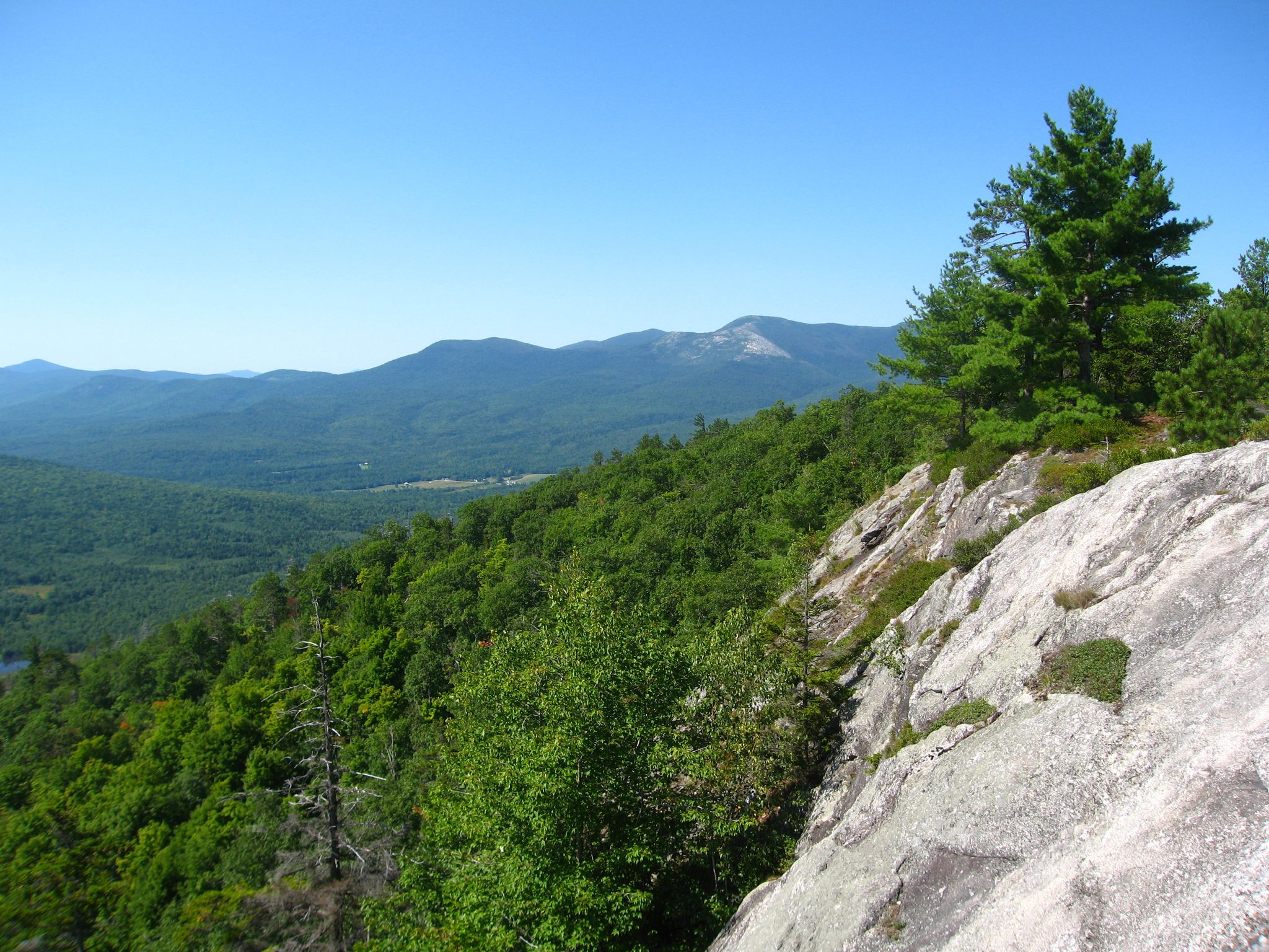 Sunny mountain vista with rocky foreground, dense green forest, and distant peaks. - Evans Notch and Blueberry Mountain Loop Trail