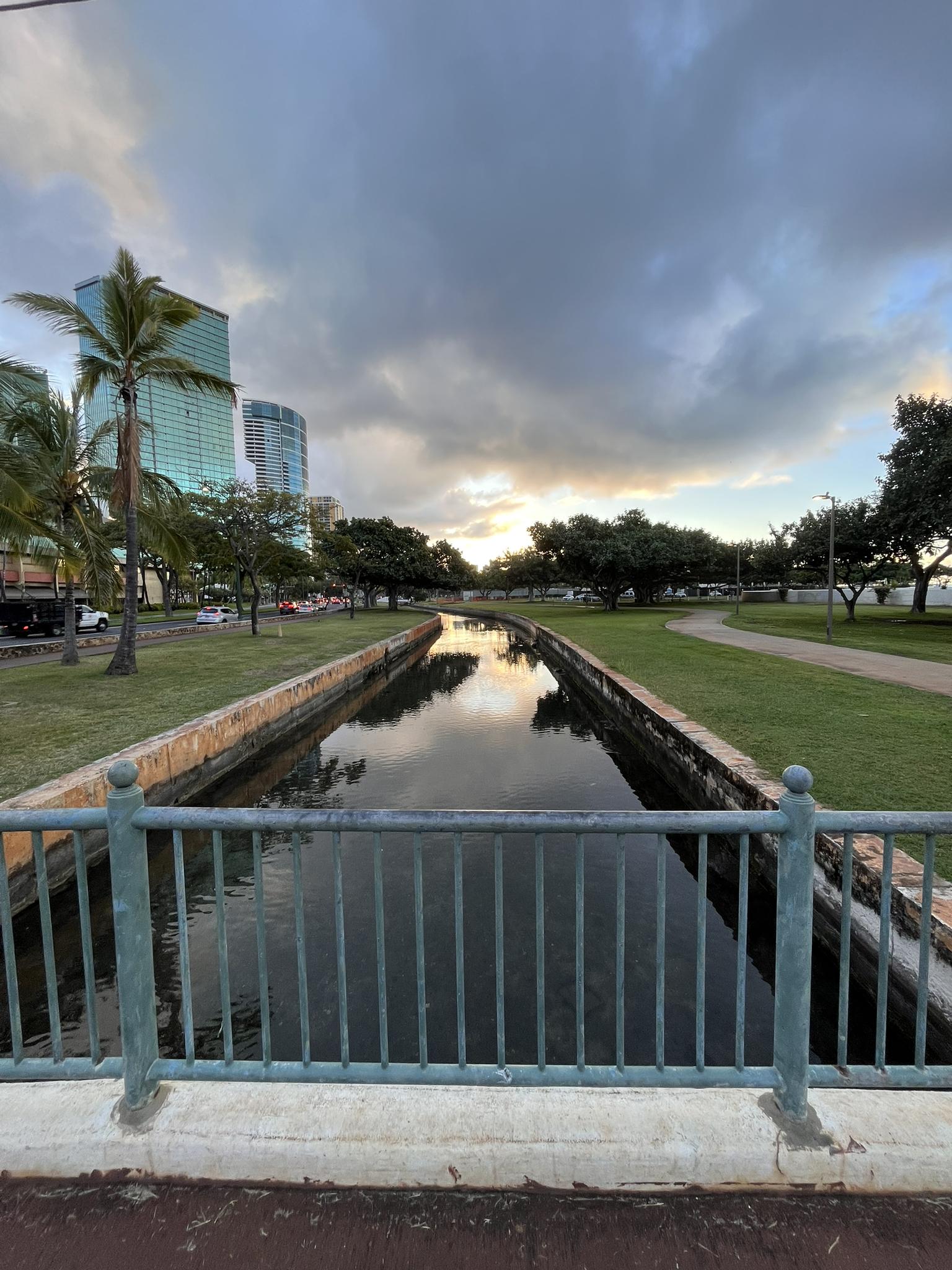 A narrow canal with grassy banks and city buildings under an overcast sunset sky.