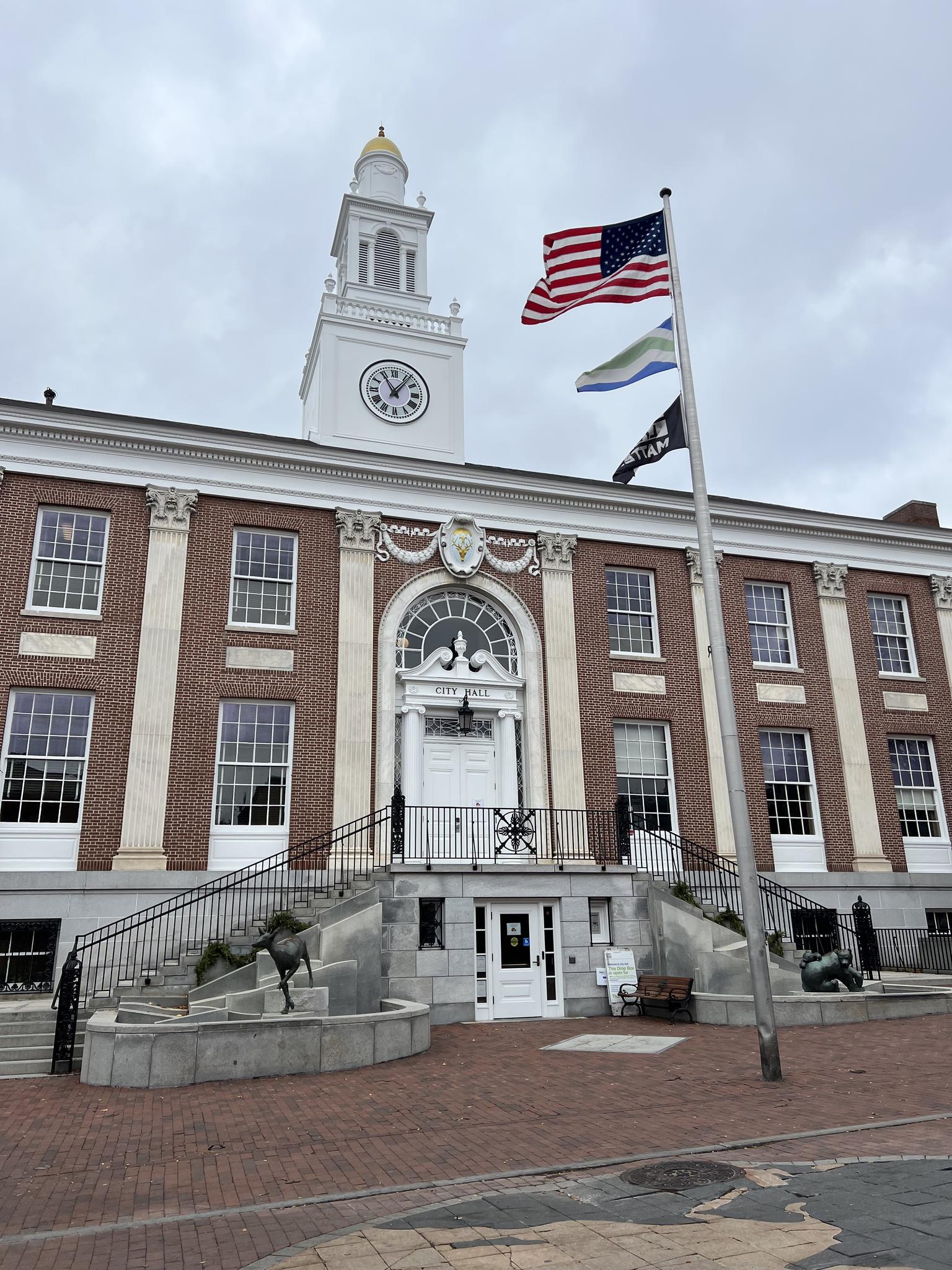 Large brick building with a white clock tower and flags under an overcast sky.