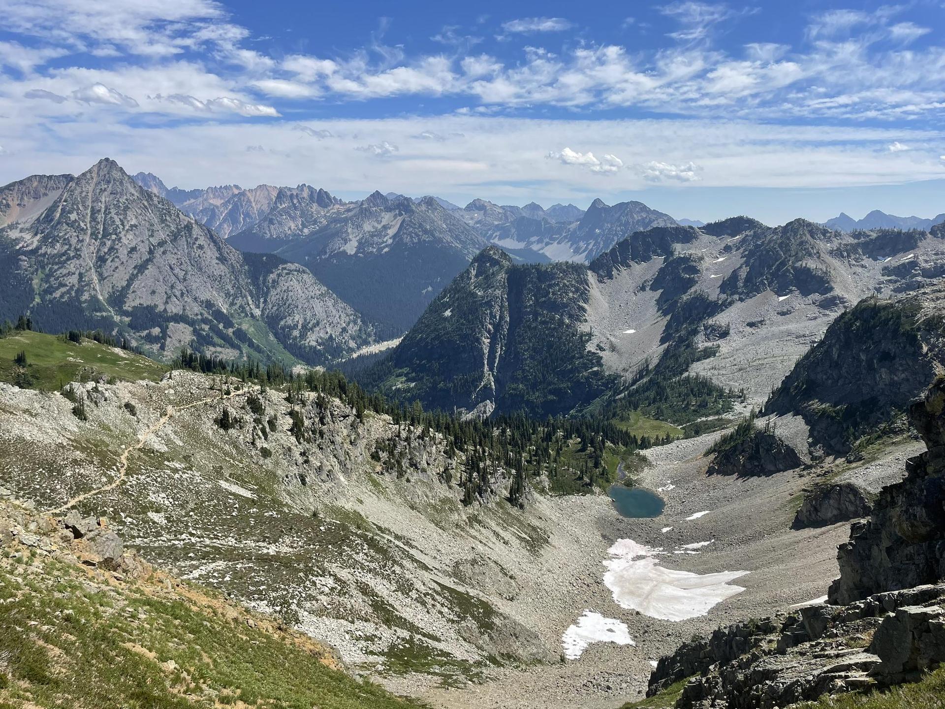 Vast mountain valley with a small lake, patches of snow, and distant peaks.