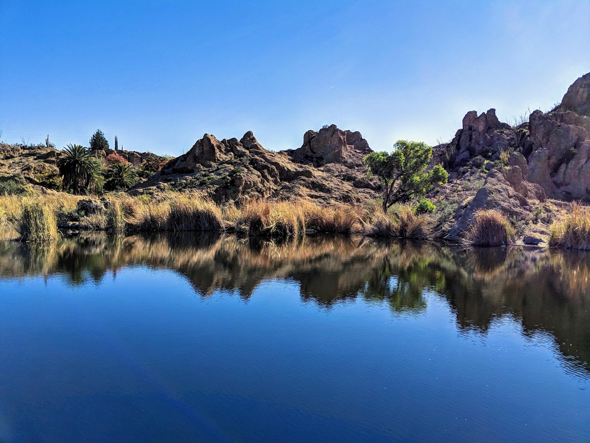 A calm lake perfectly reflecting rocky hills and dry grass under a blue sky.