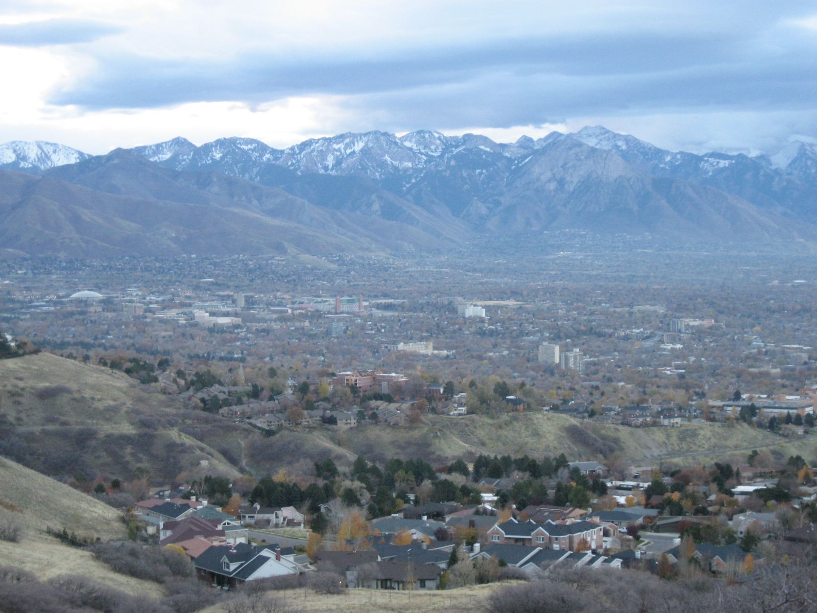 Overcast view of a city and houses nestled below large snow-capped mountains. - Meridian Peak