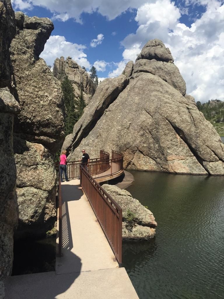 People on a boardwalk next to a lake, surrounded by large rock formations. - Sylvan Lake Shore Trail