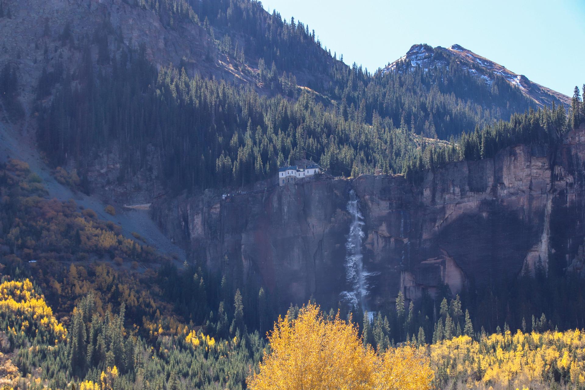 White building on a cliff above a waterfall, surrounded by autumn trees.