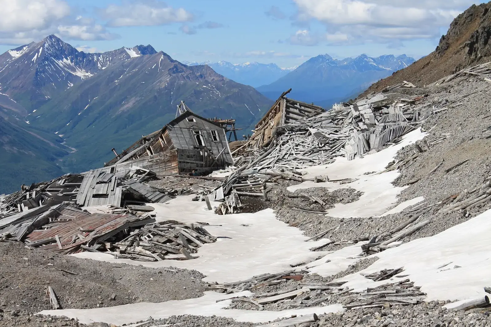 Dilapidated wooden buildings on a rocky mountain slope with snow patches.