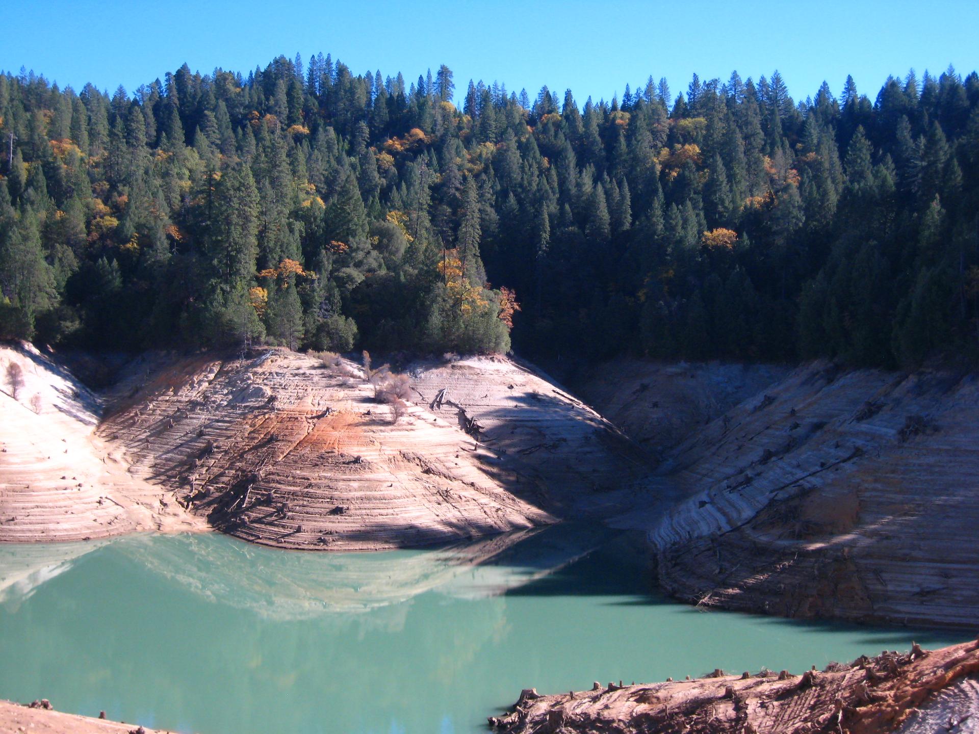Greenish water in a reservoir with terraced, exposed banks and forested hills.