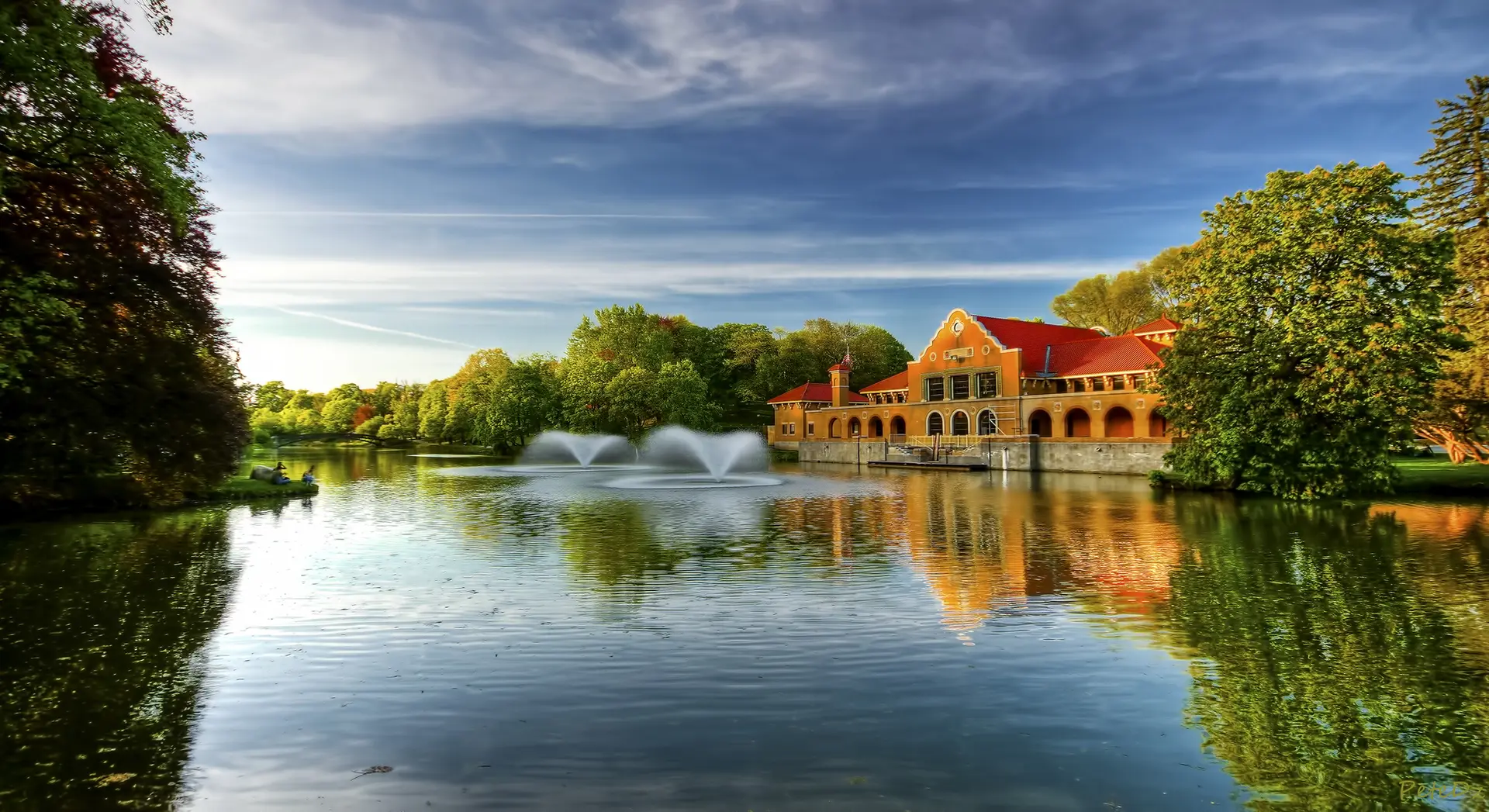 Scenic lake with fountains, a large building, and lush green trees under a bright sky.