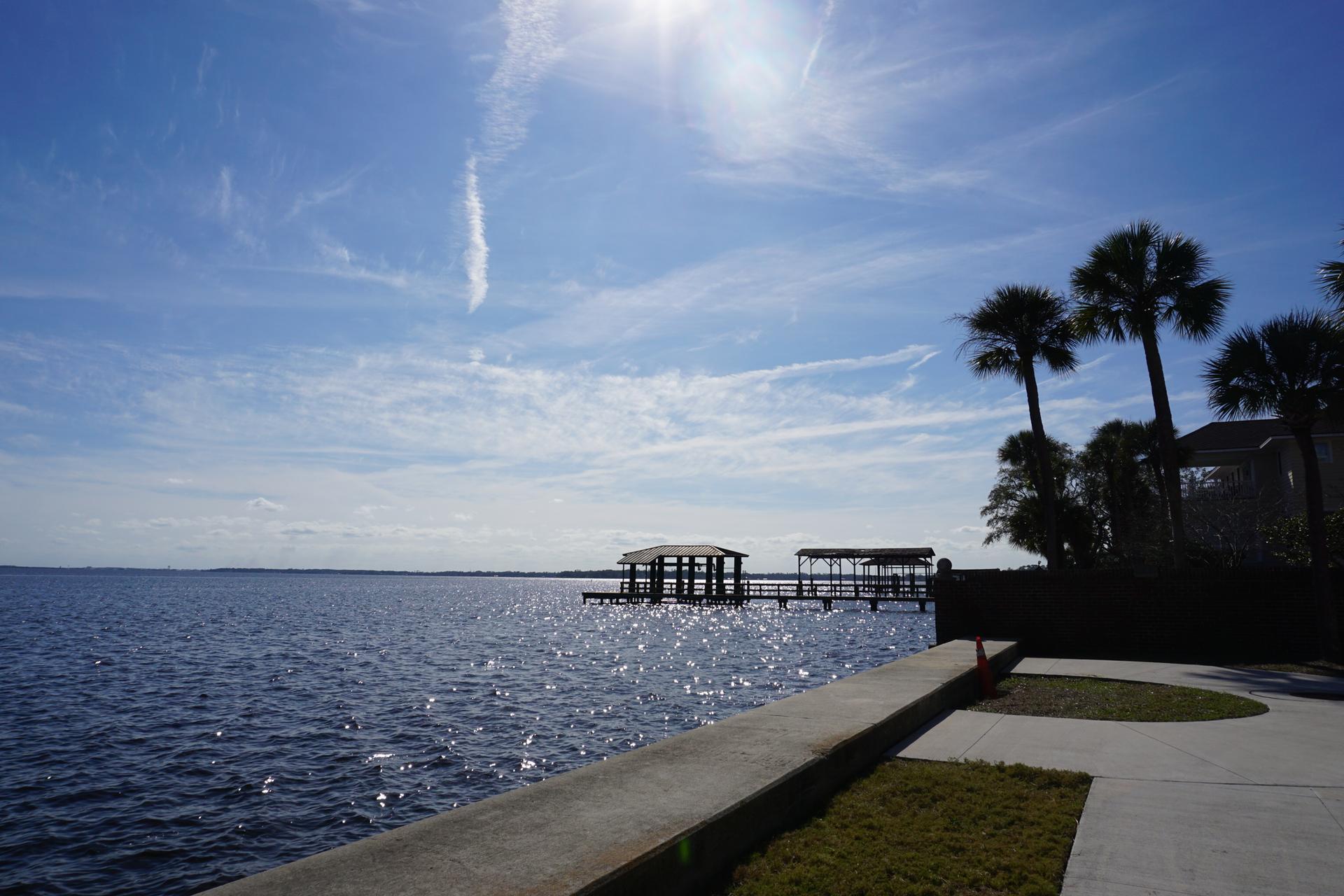 Bright sun over calm water with a distant pier and palm trees.