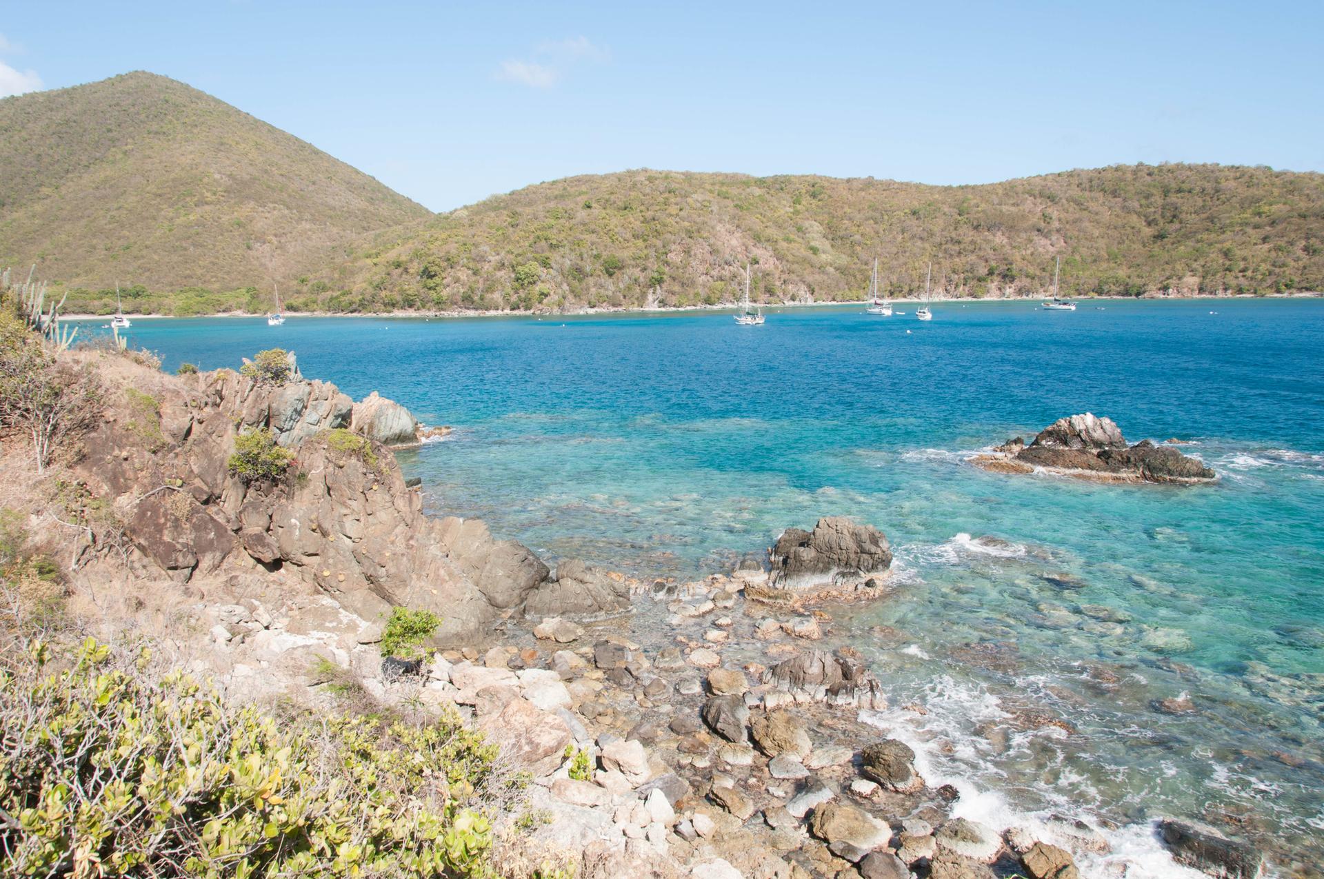 Bright sunny view of clear blue water, rocky coast, and distant sailboats.