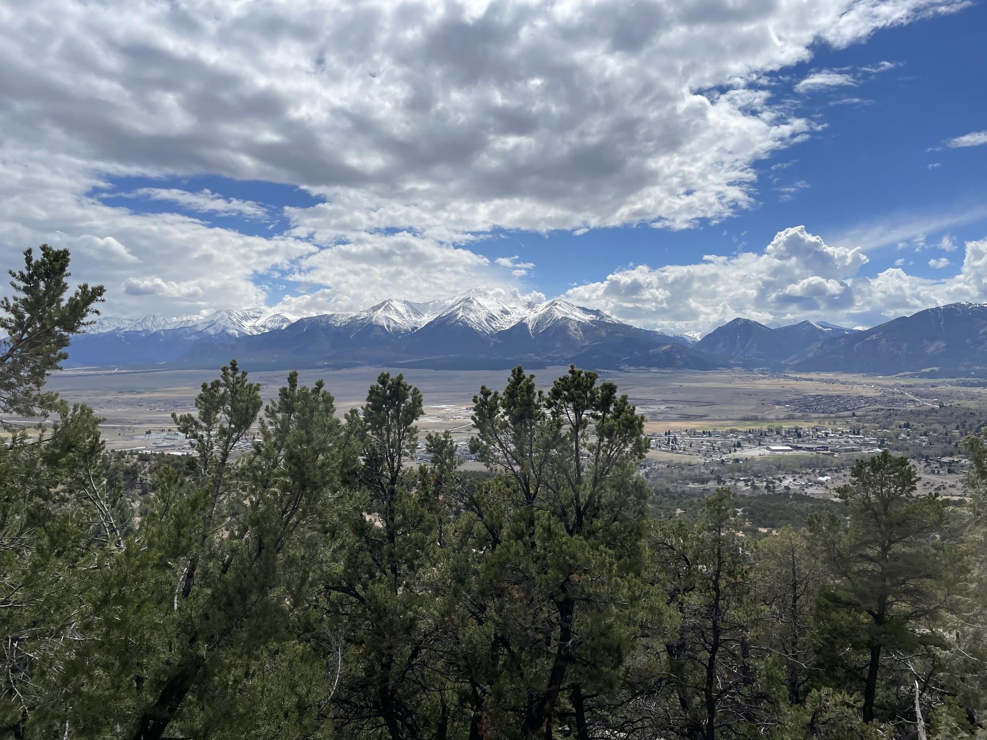 Sunny view of snow-capped mountains and a valley with a town, framed by trees.