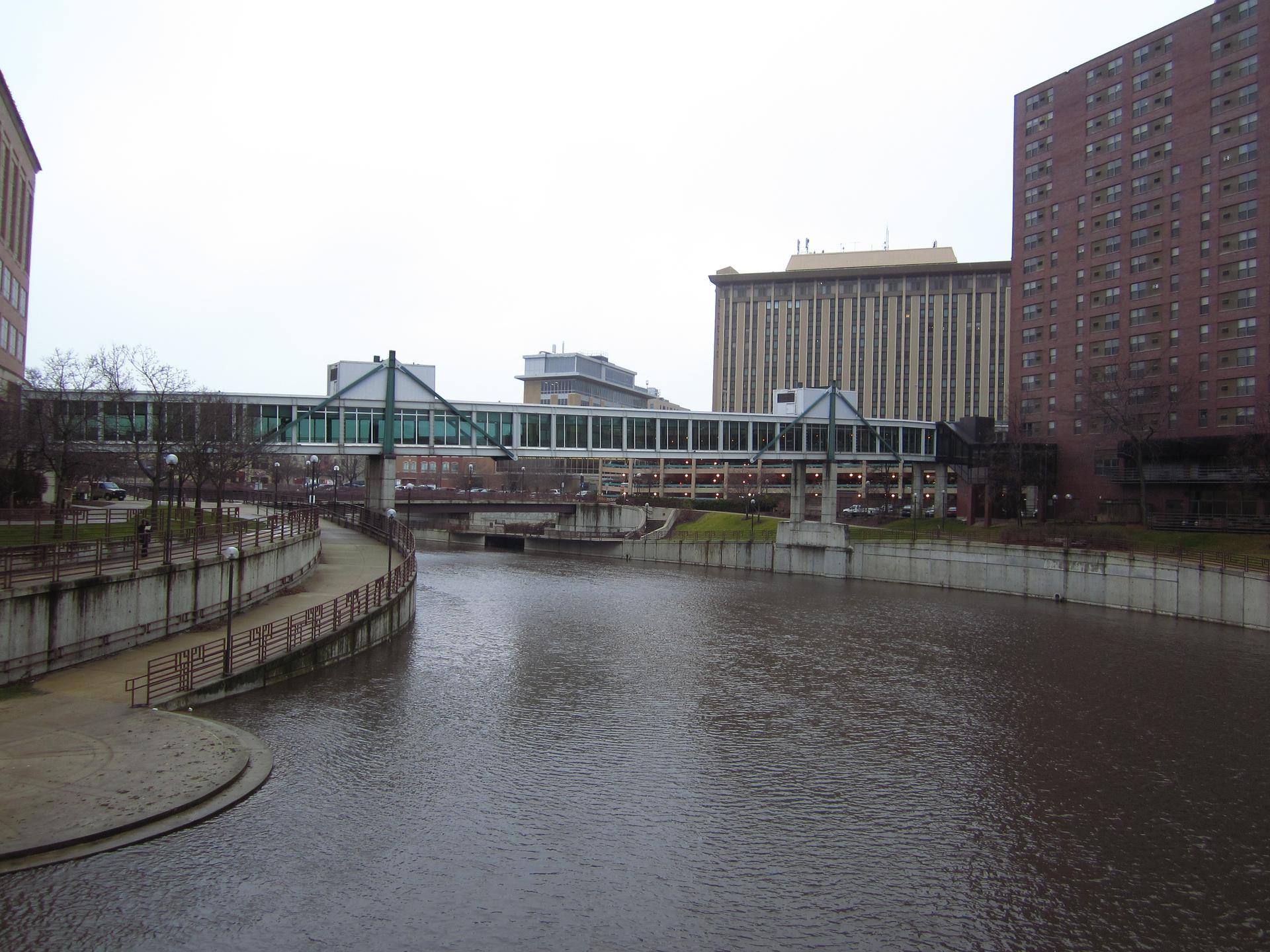 Wide river flowing through a city with buildings and an enclosed pedestrian bridge.