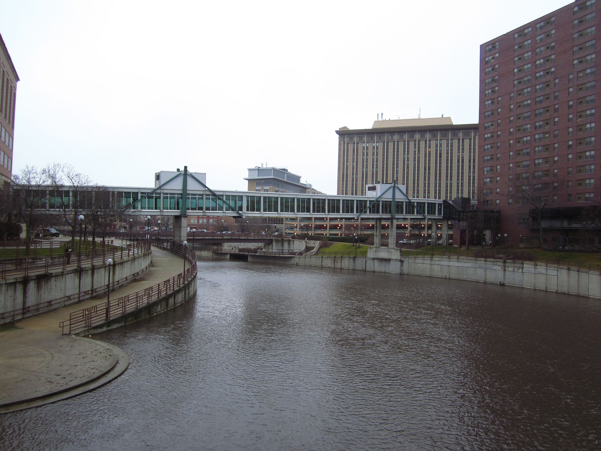 Wide river flowing through a city with buildings and an enclosed pedestrian bridge. - Zumbro River Trail